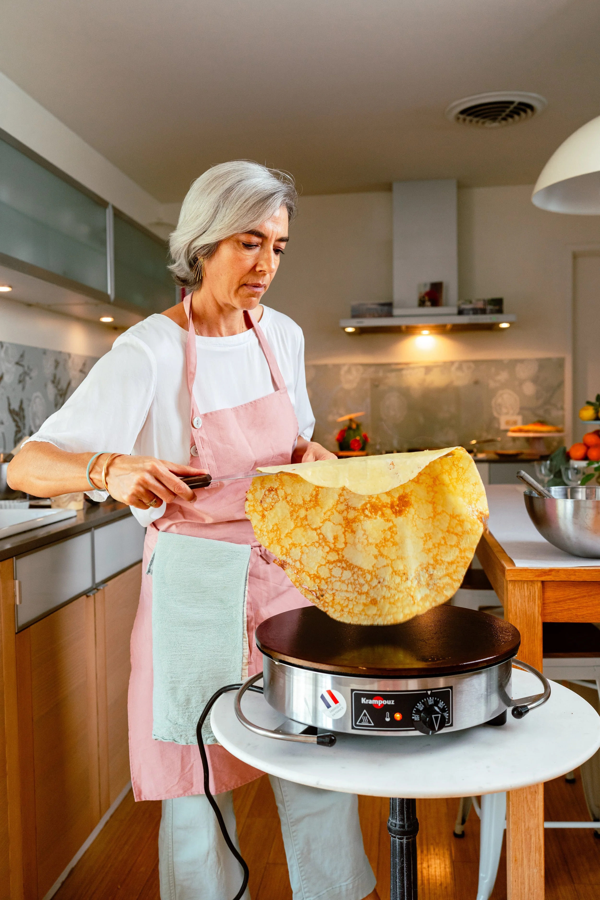 A woman in a pink apron is making crêpes using a large spatula on an electric griddle in a modern kitchen.