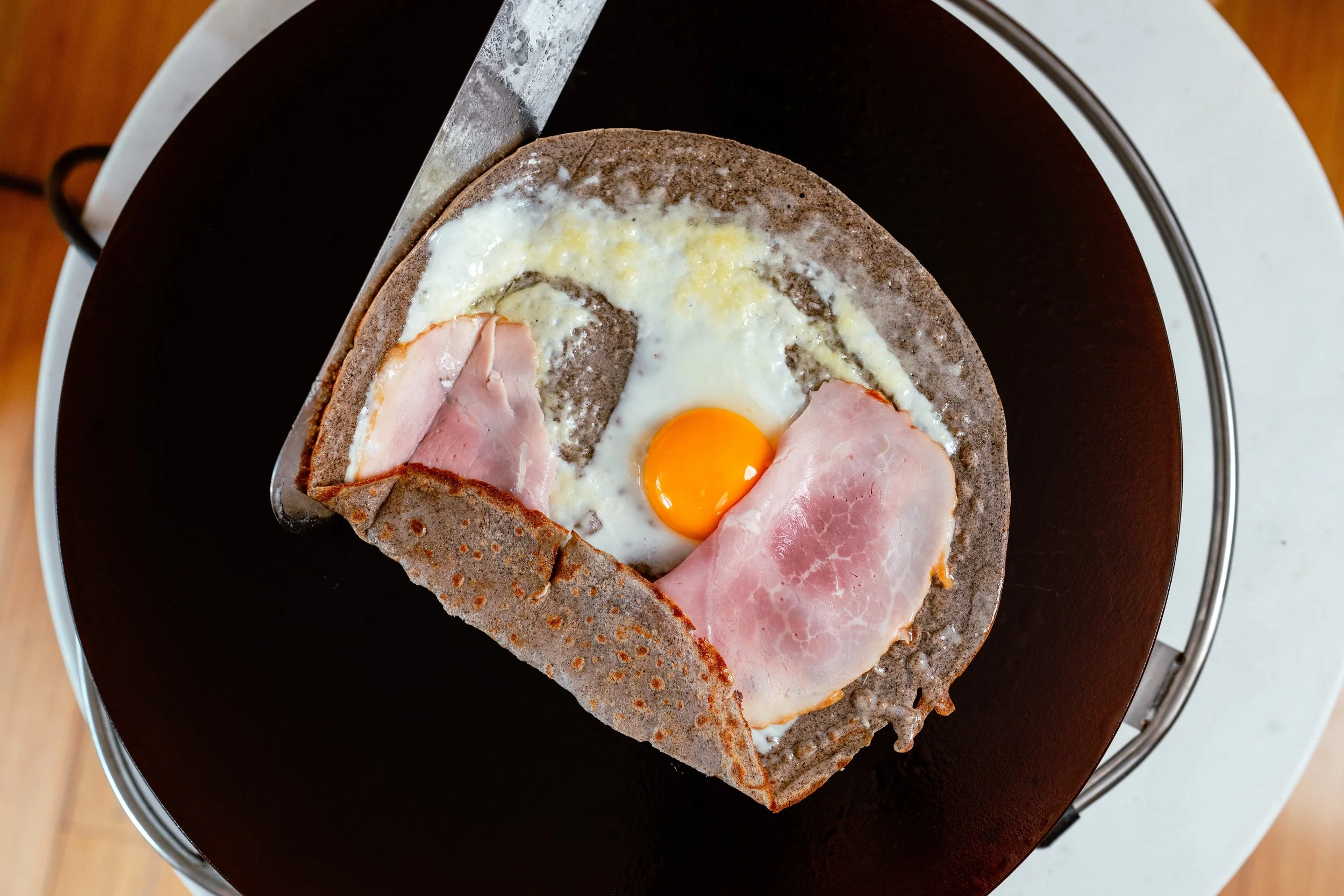 A buckwheat crepe with an egg in the center and slices of ham on the sides, cooking on a professional crepe griddle.