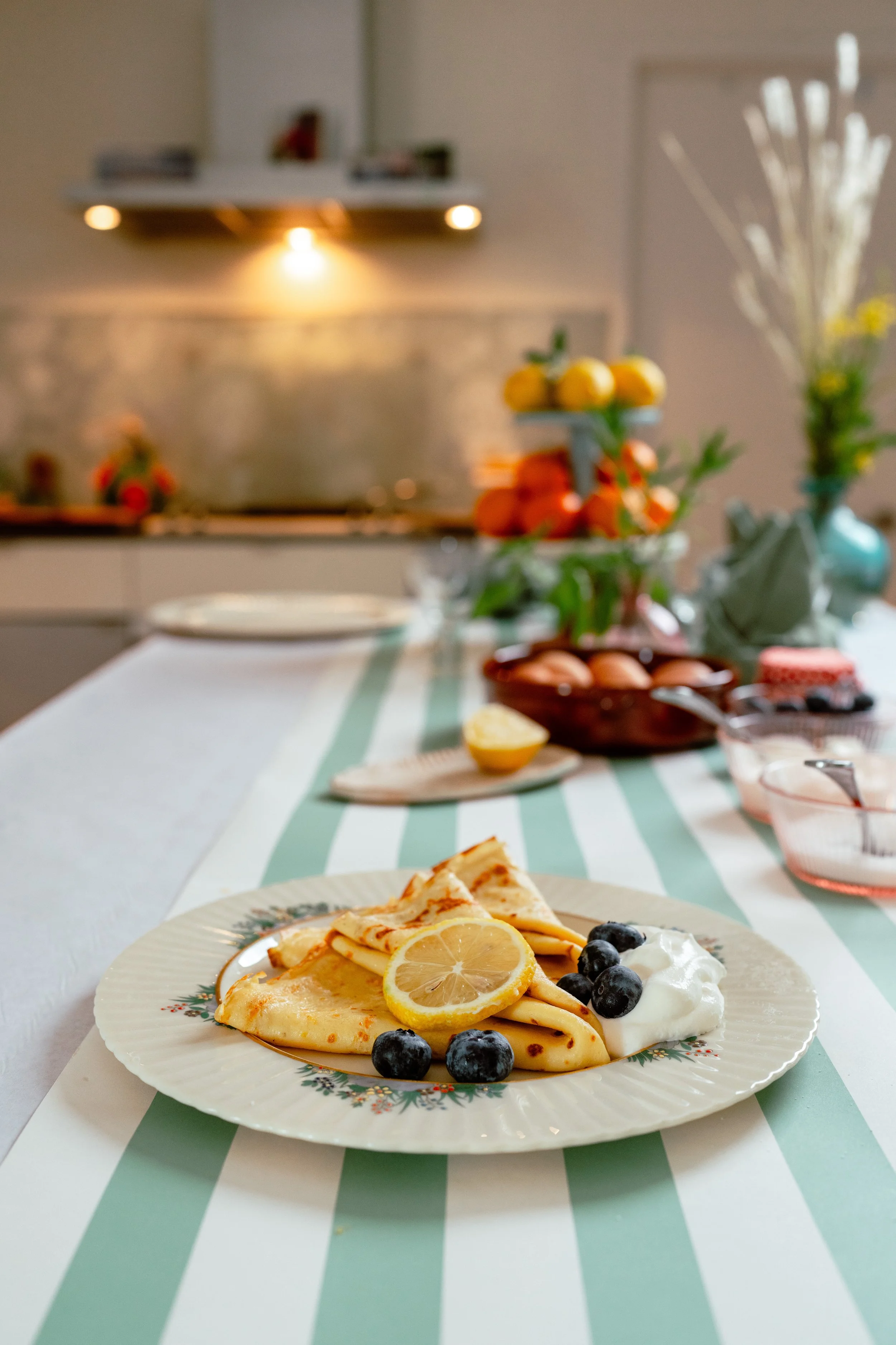 A breakfast plate with crepes topped with lemon slices and blueberries, served with whipped cream, on a table with a striped tablecloth. In the background, a kitchen with a stove, fruit, and flowers.