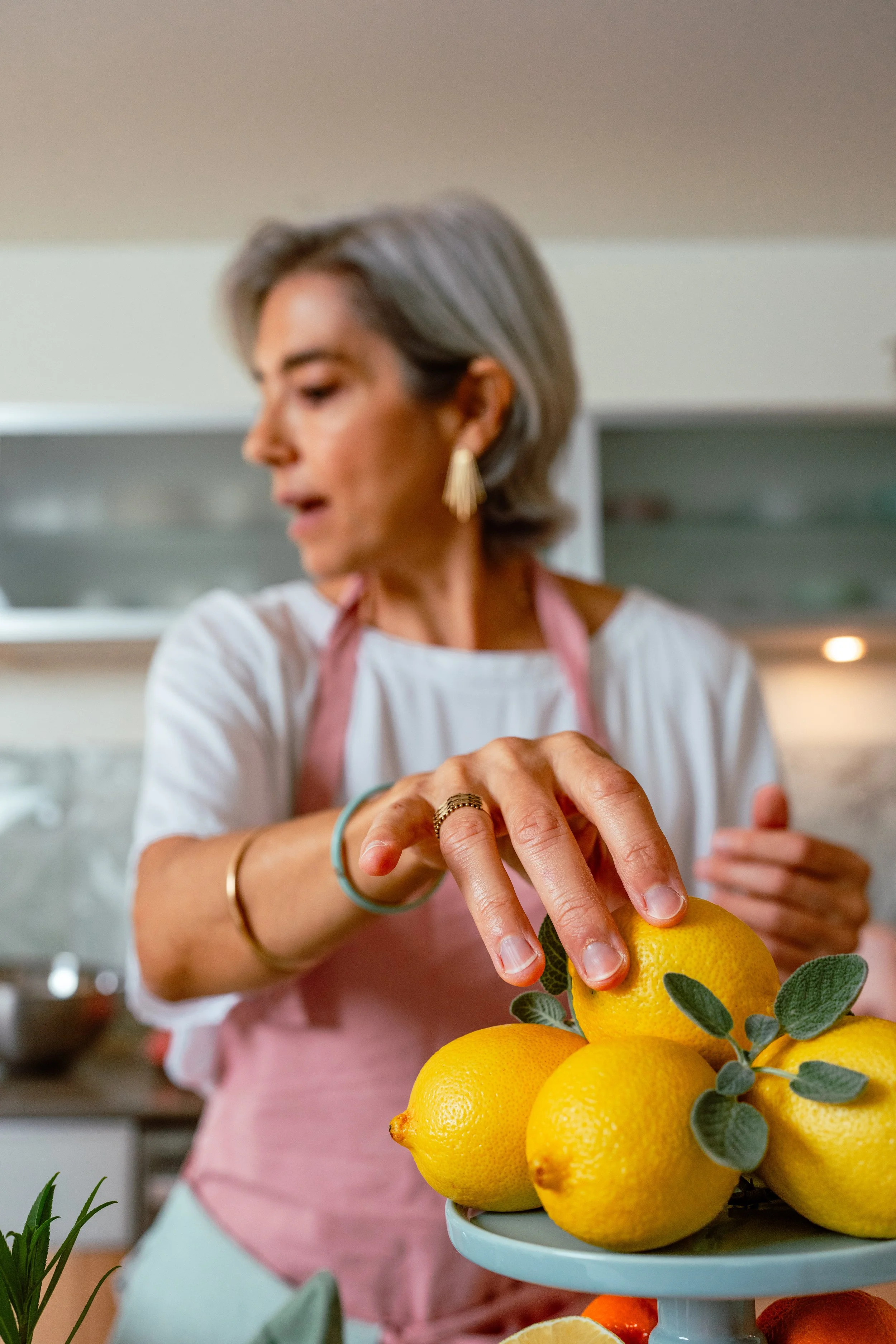 A woman with short gray hair and gold earrings, wearing a pink apron, is picking a lemon from a plate of lemons in a kitchen.