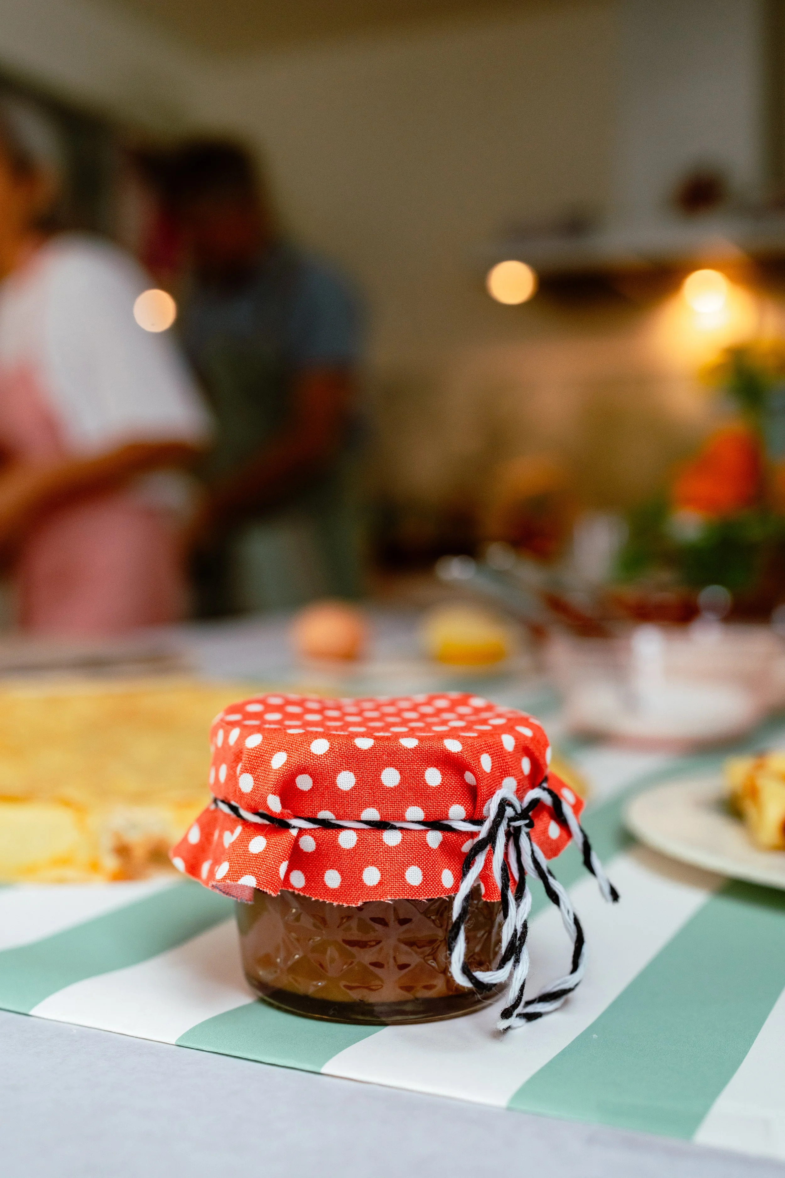 Close-up of a jar with a red polka dot cloth cover tied with black and white string, placed on a table with other blurred food items and people in the background.