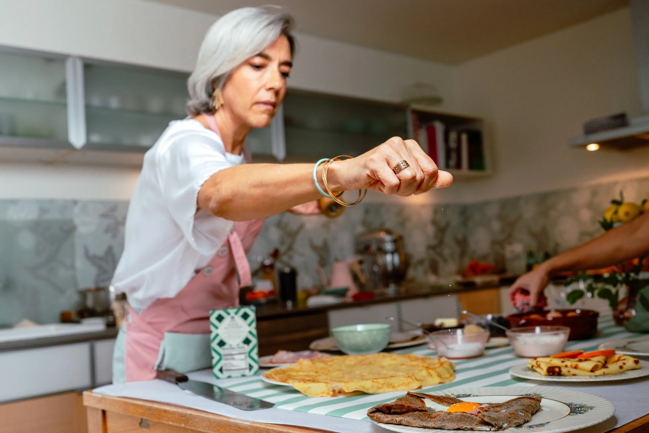 A woman in a kitchen preparing food, with a buckwheat crepe, and other dishes on the table.