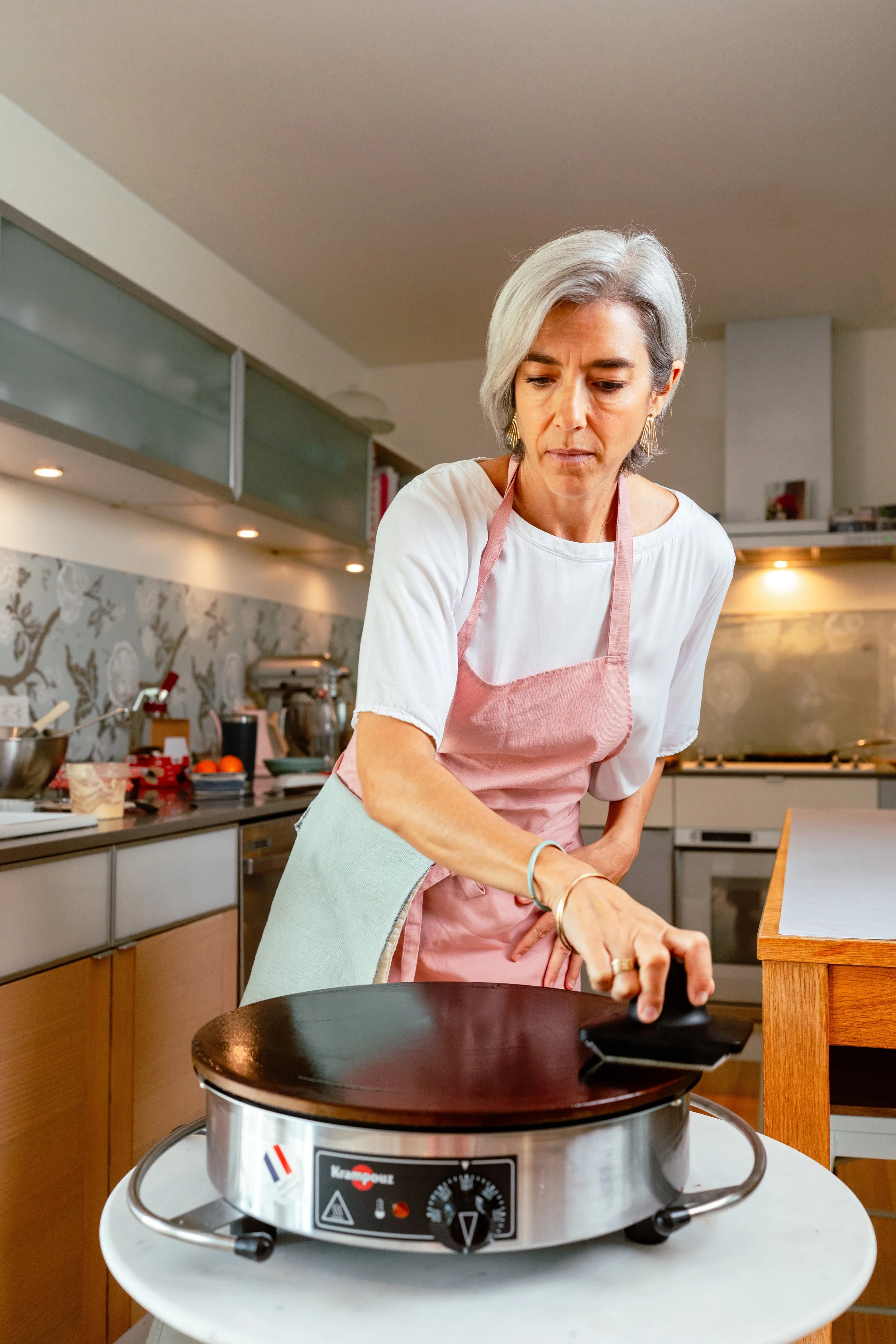 A woman wearing a white shirt and pink apron is cleaning an electric crepe maker on a white table in a modern kitchen.