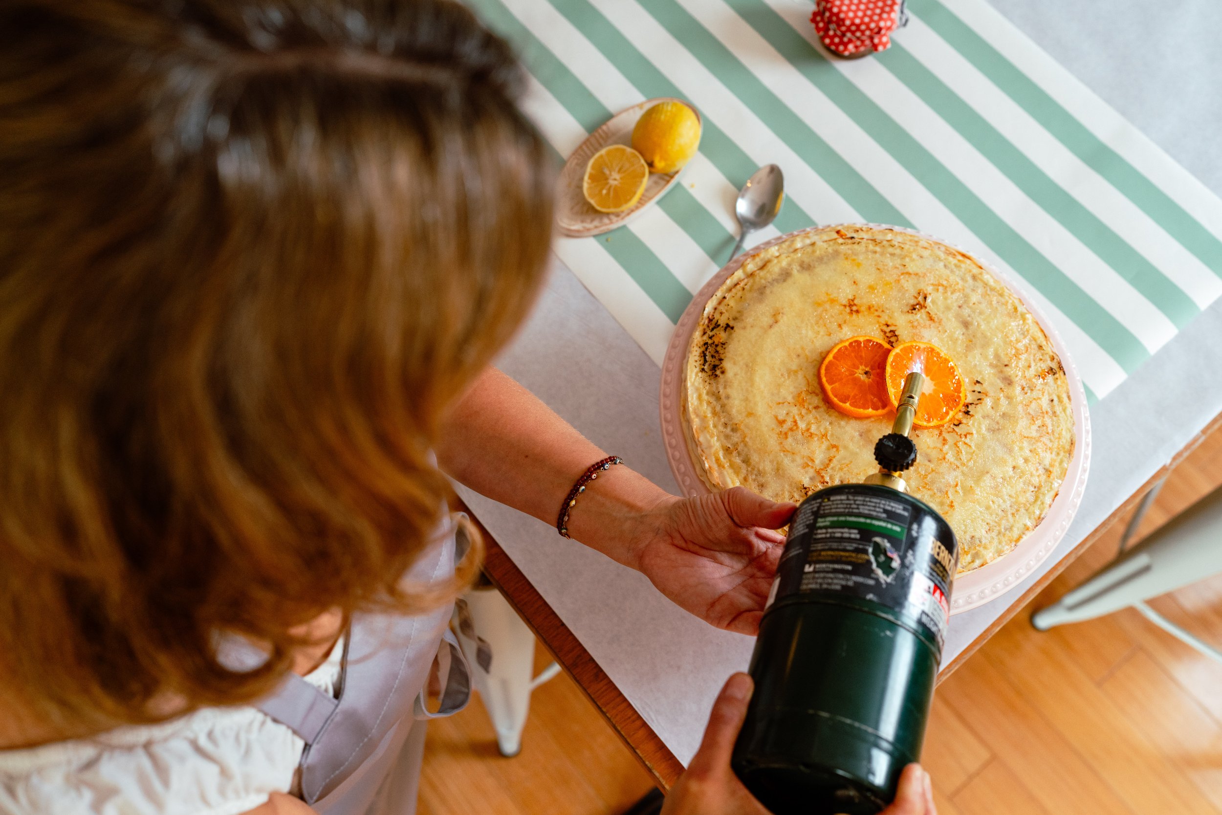 A person is bruleeing a crepe cake topped with orange slices with a culinary torch. The cake is on a pink plate on a table covered with a white and green striped tablecloth. Nearby, there is a wooden plate with a lemon, lemon half, and a spoon.