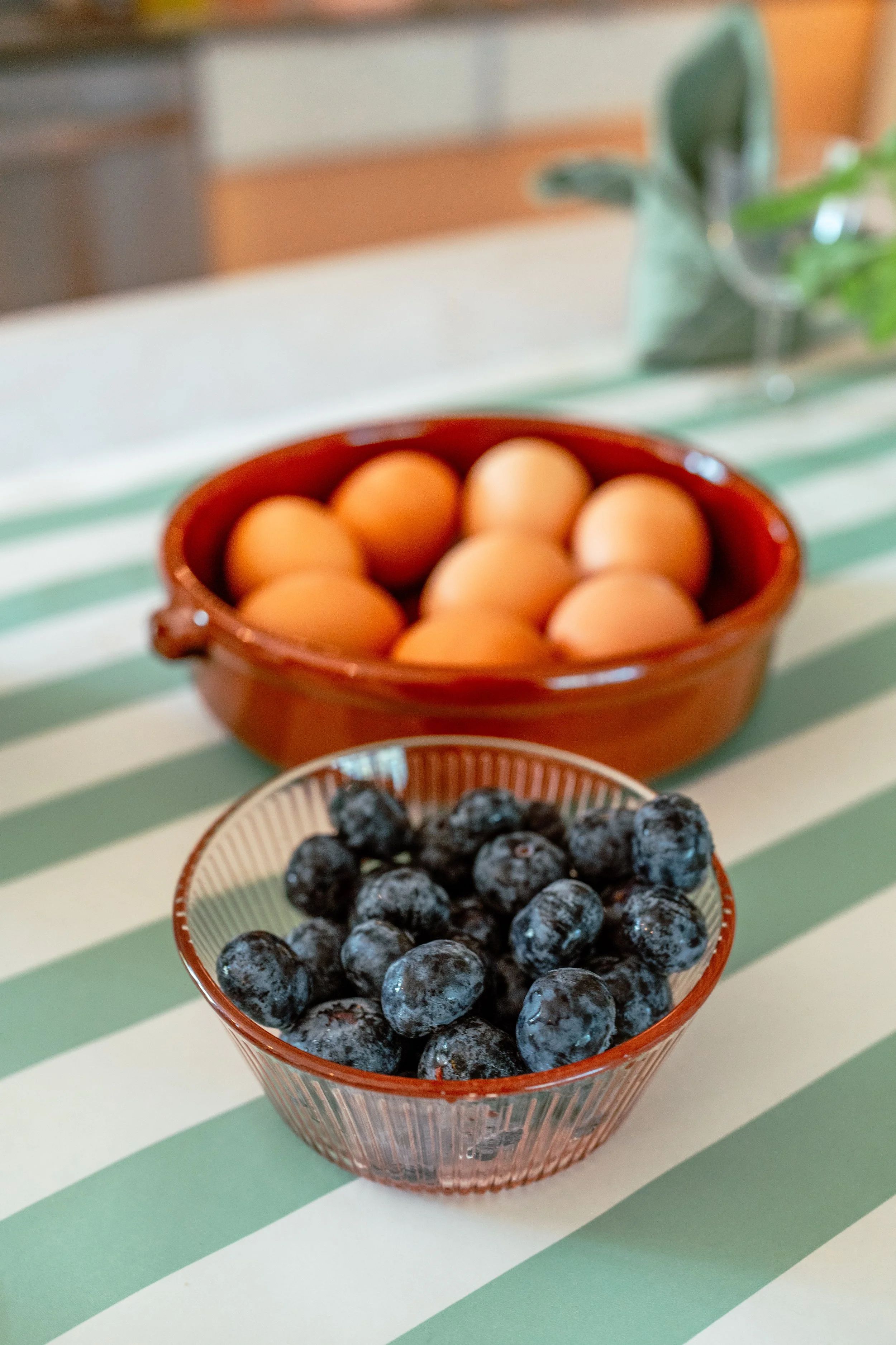 A small pink bowl filled with blueberries and a larger terracotta bowl filled with eggs, placed on a striped tablecloth.