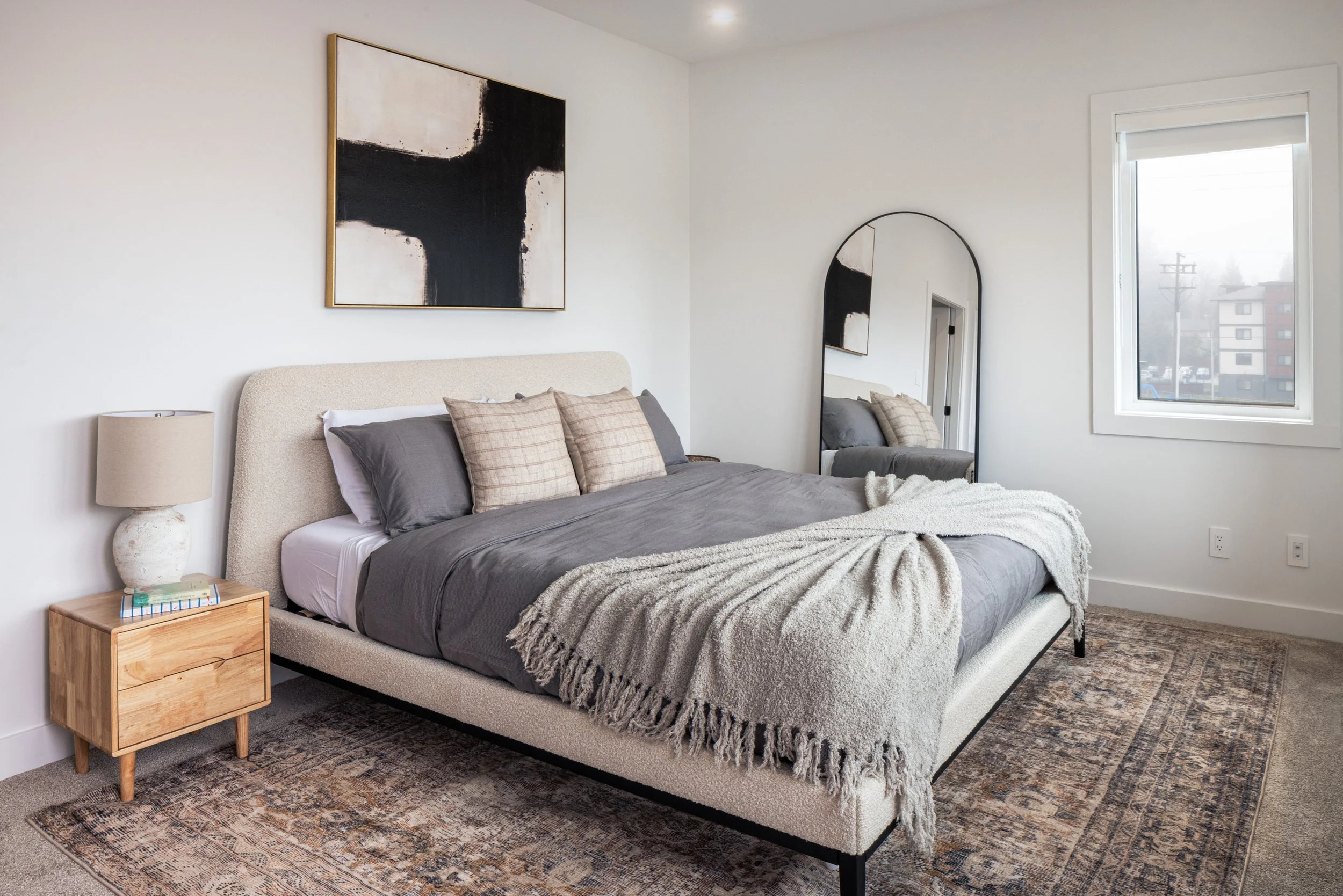 Modern bedroom with a beige upholstered bed, gray bedding, plaid pillows, a nightstand with a lamp and books, a large abstract black and white wall art, a tall arched mirror, a window showing an outdoor view, and a patterned rug.