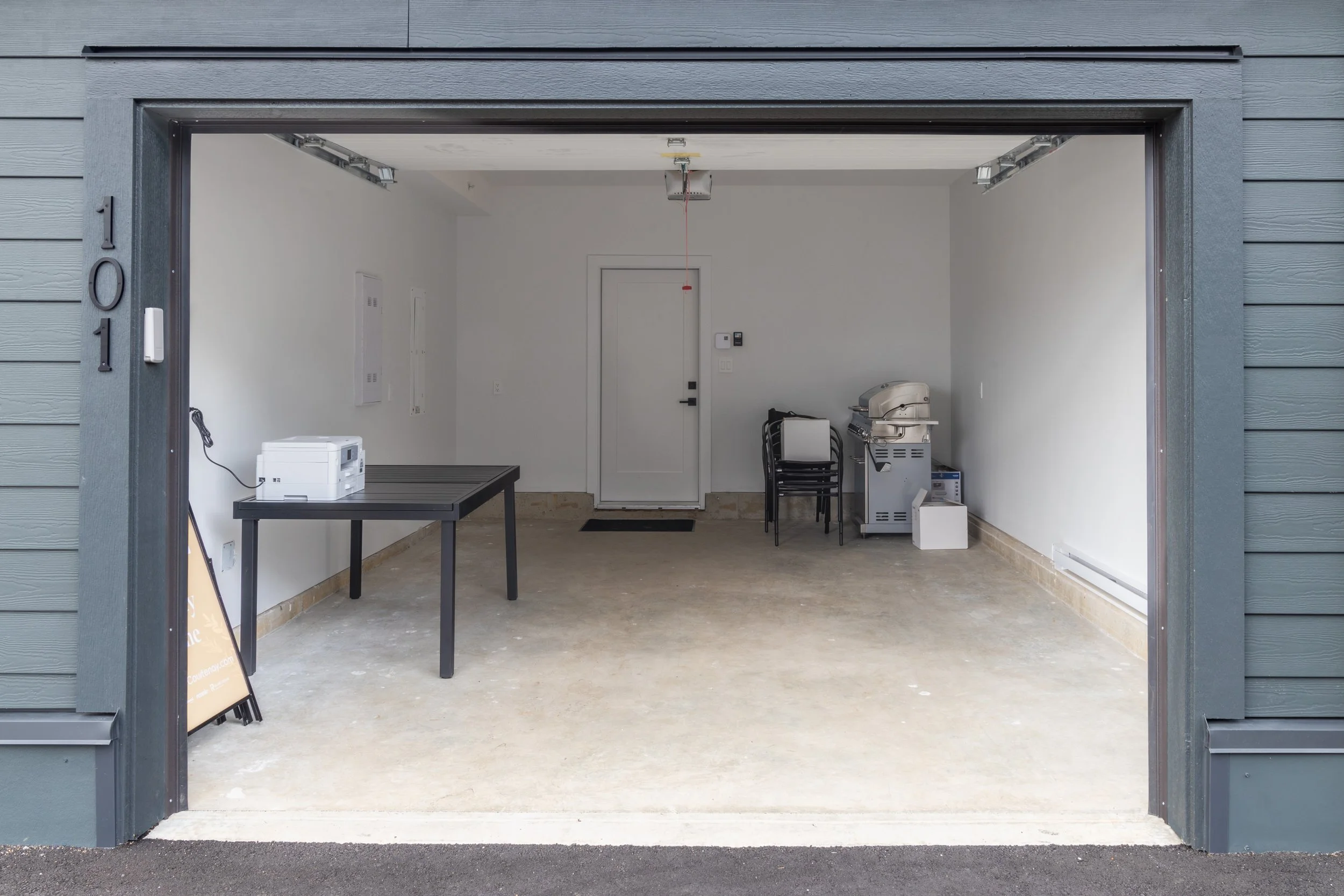 Empty garage with a table, a printer, stacked chairs, a grill, and a cardboard box, with a door and some electrical panels on the back wall.