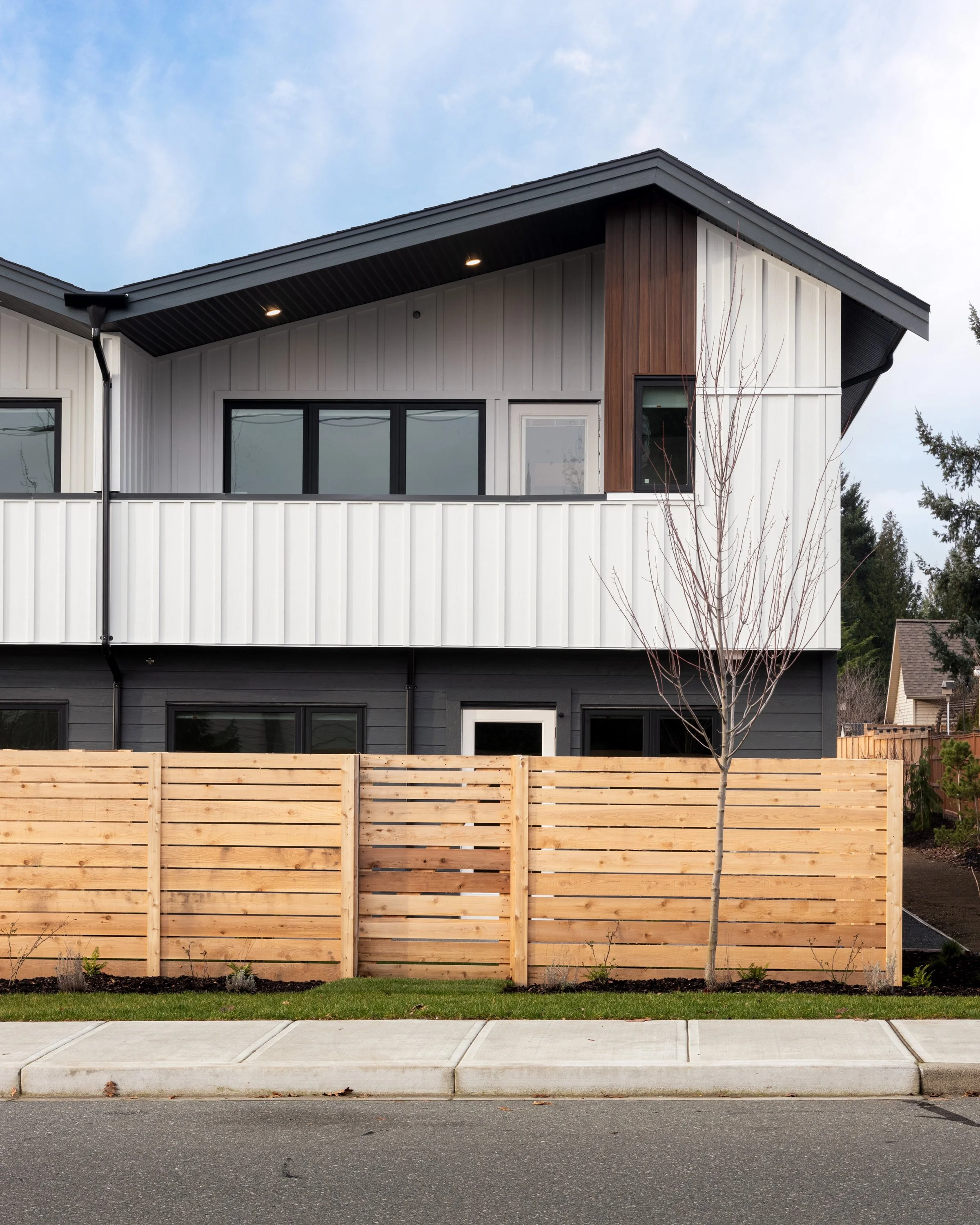 Modern two-story house with white and dark gray siding, small tree in front, and a wooden fence along the sidewalk.