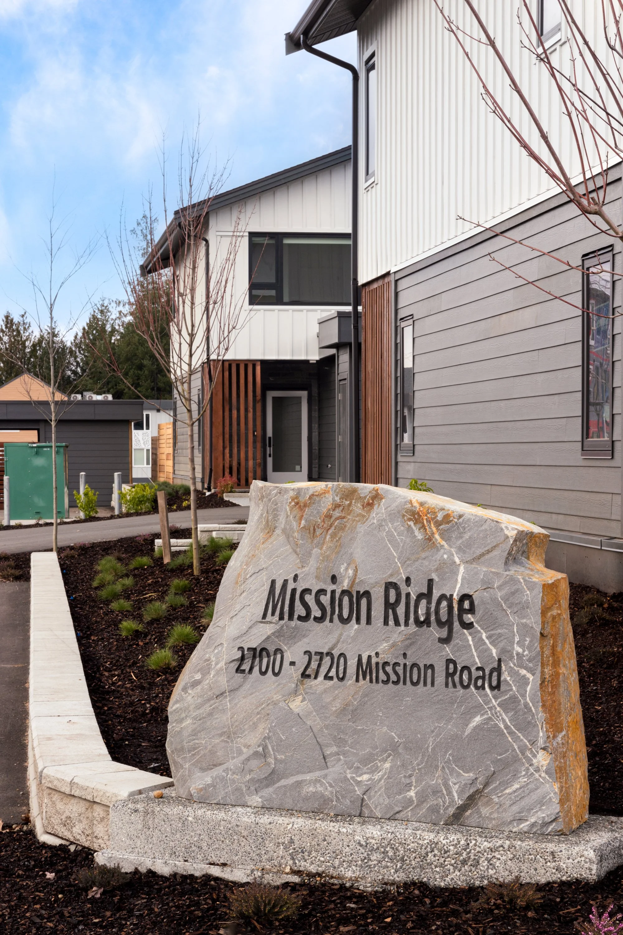 A large decorative stone at the entrance of a residential neighborhood reading "Mission Ridge, 2700 - 2720 Mission Road," with modern houses and trees in the background.
