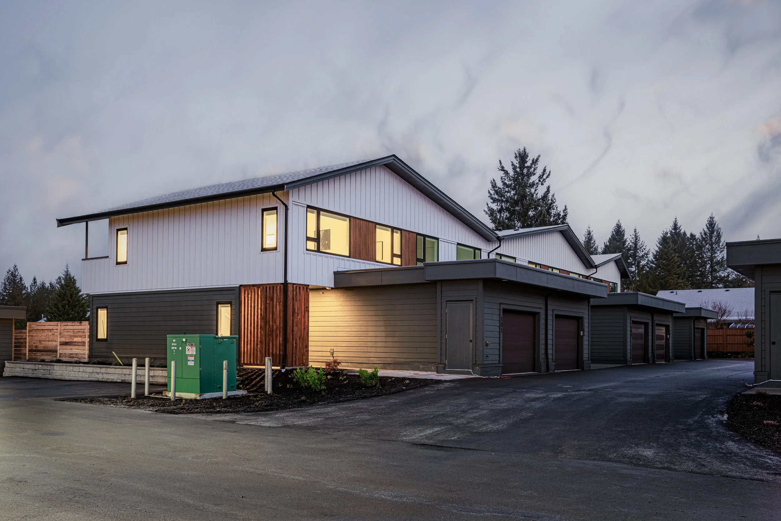 Multi-unit residential building with garage units, exterior lighting on at dusk, surrounded by trees, driveway, and fencing.
