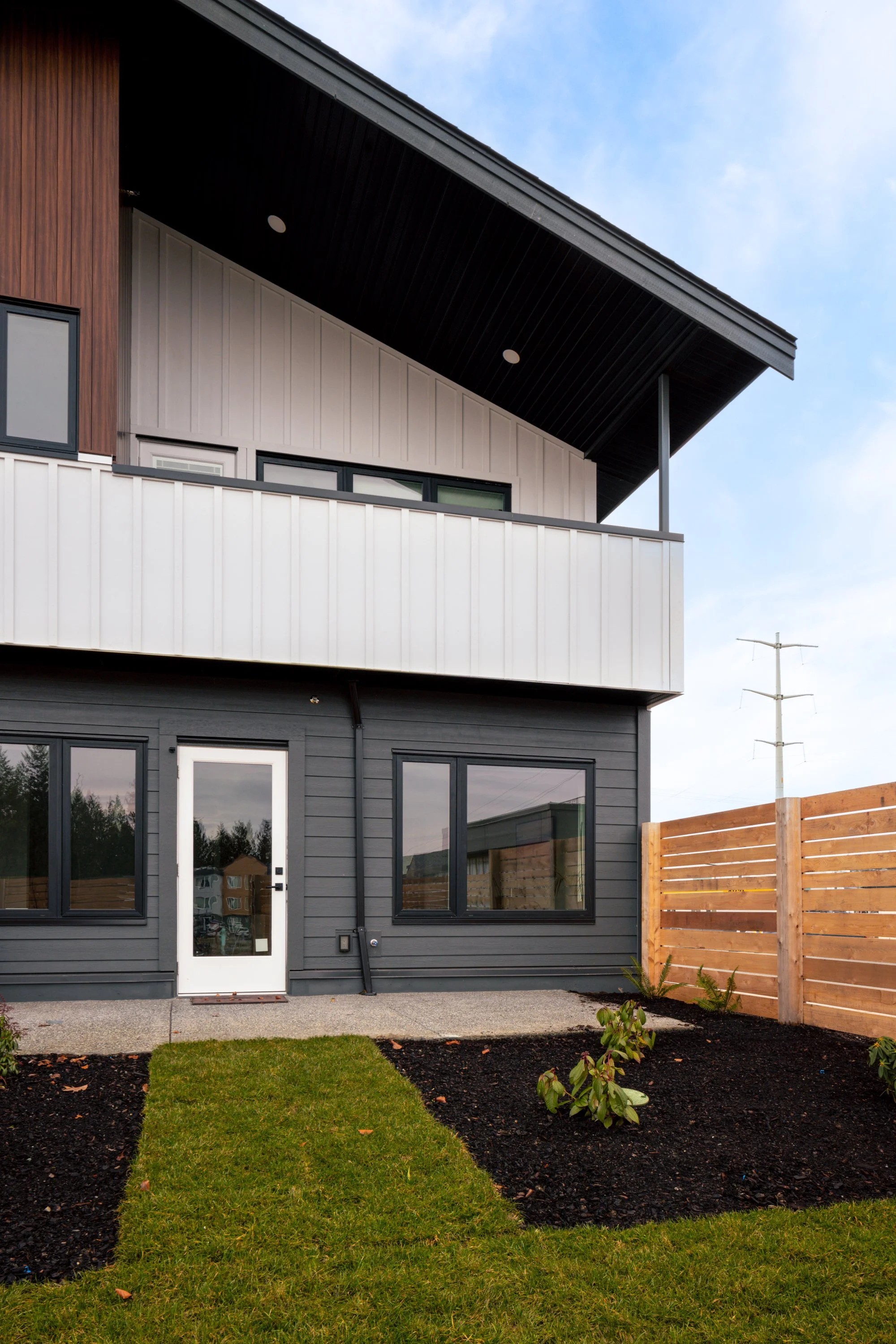Exterior view of a modern two-story house with dark gray siding, a wooden fence, and a small landscaped yard with grass and young plants.