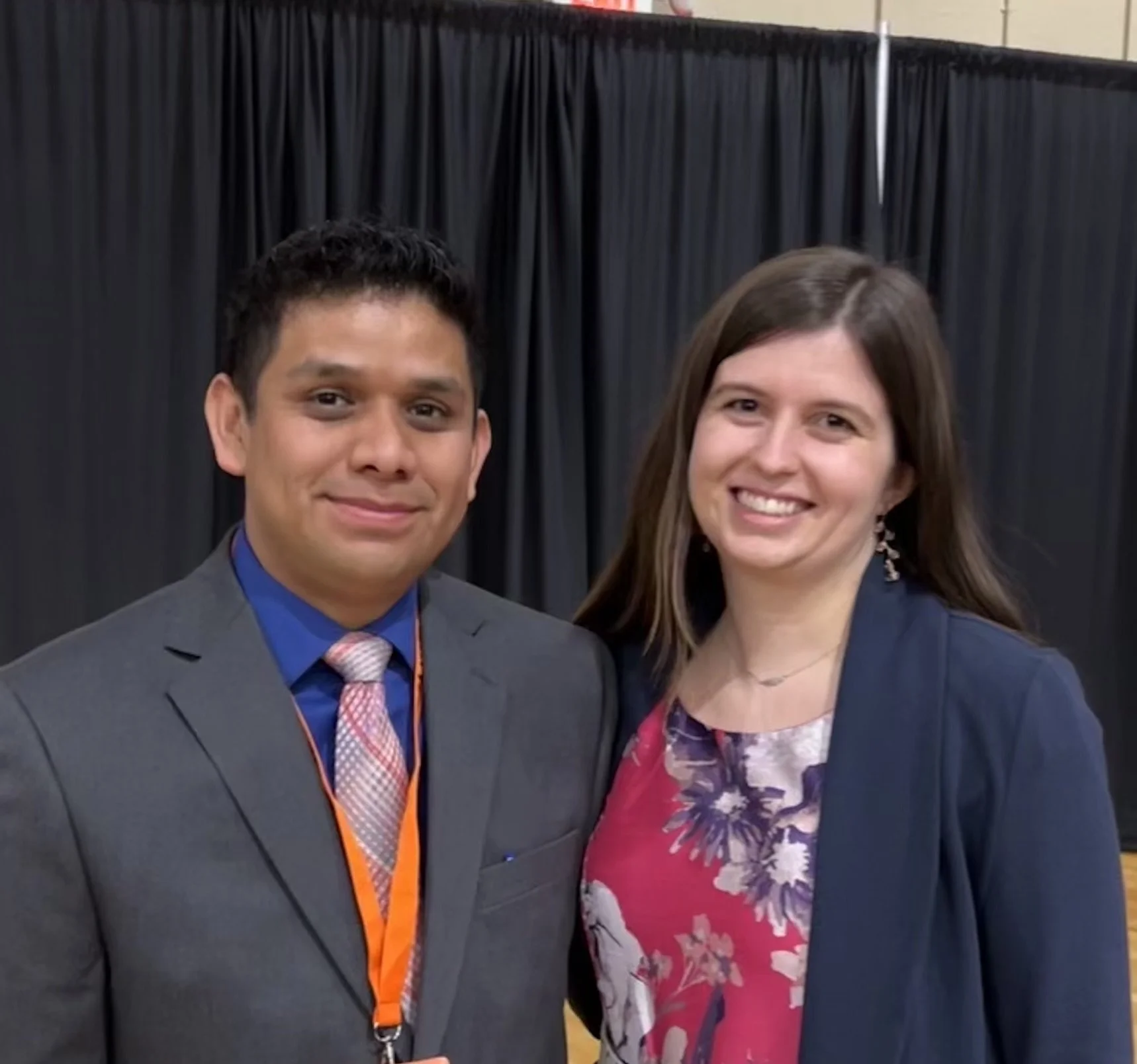 A man in a suit and a woman in a floral top and dark blazer standing together and smiling at an indoor event with a black curtain backdrop.