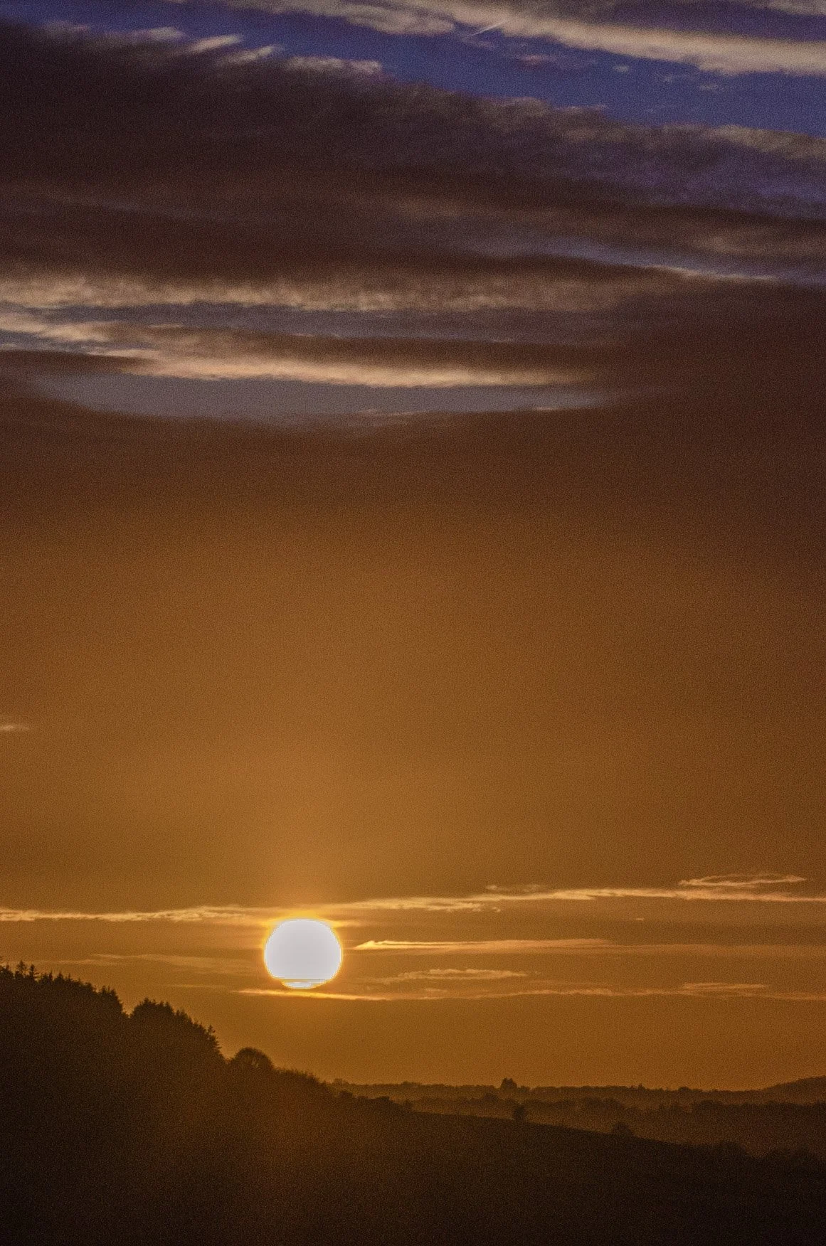 Sunset over a hilly landscape with dark silhouettes of trees and scattered clouds in the sky.