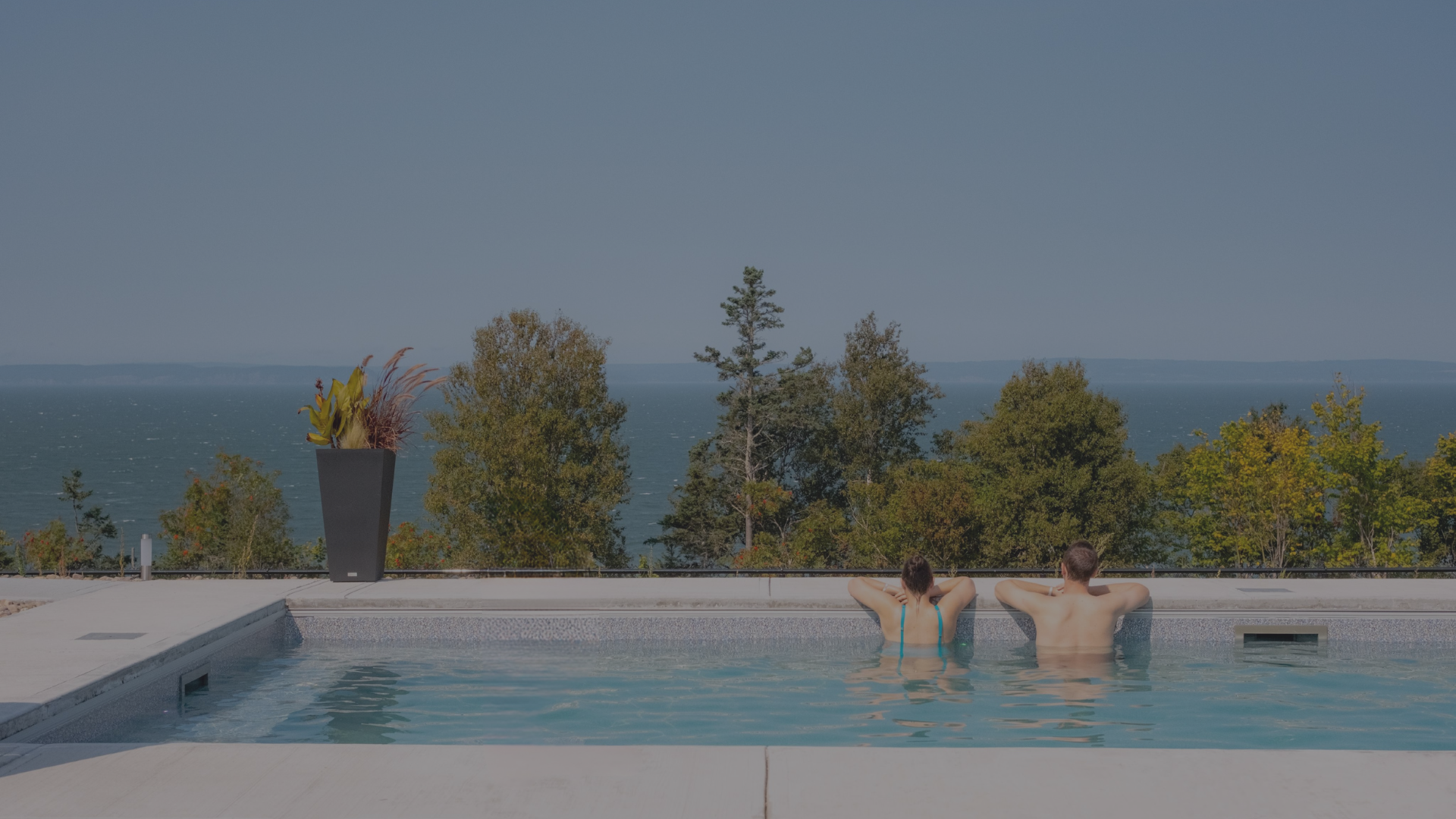 Two people are relaxing in an outdoor swimming pool, facing a scenic view of trees and a body of water in the distance on a sunny day.
