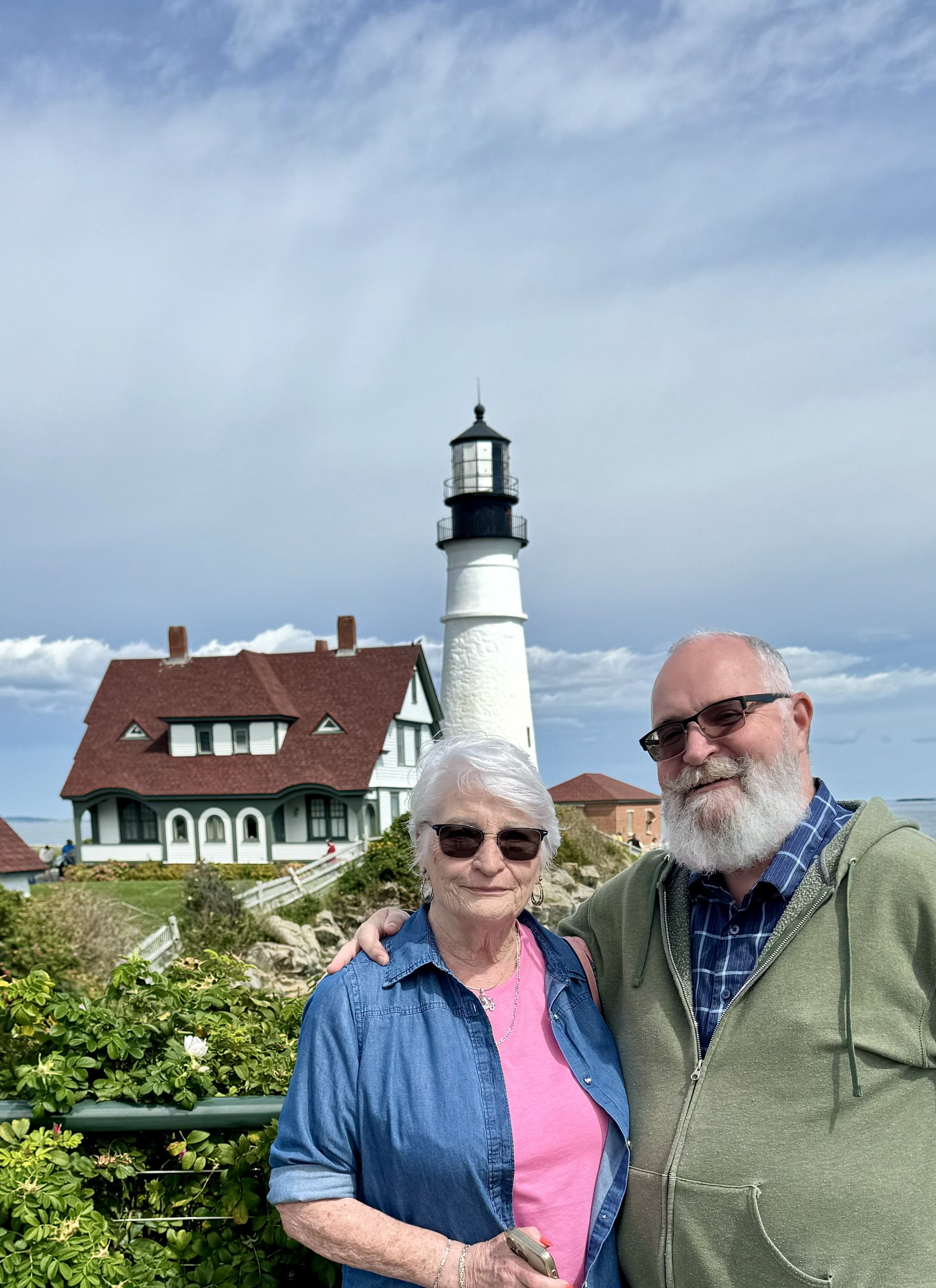 Two elderly people, a woman and a man, standing outdoors near a lighthouse, with the woman wearing glasses and a denim shirt, and the man wearing glasses and a green jacket, smiling at the camera.