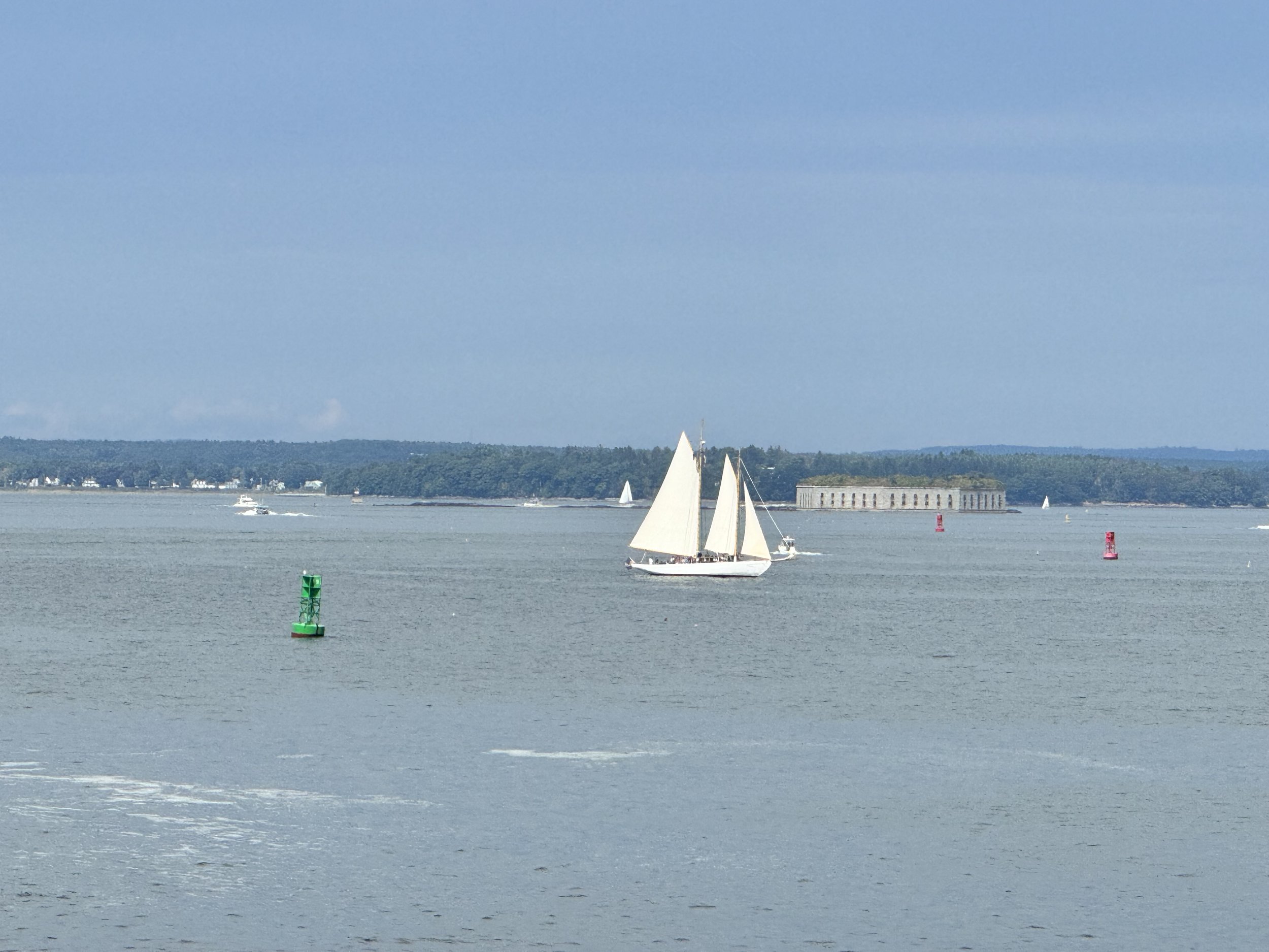 Sailboat on the water with green and red navigation buoys, distant trees, and an aged brick structure in the background.