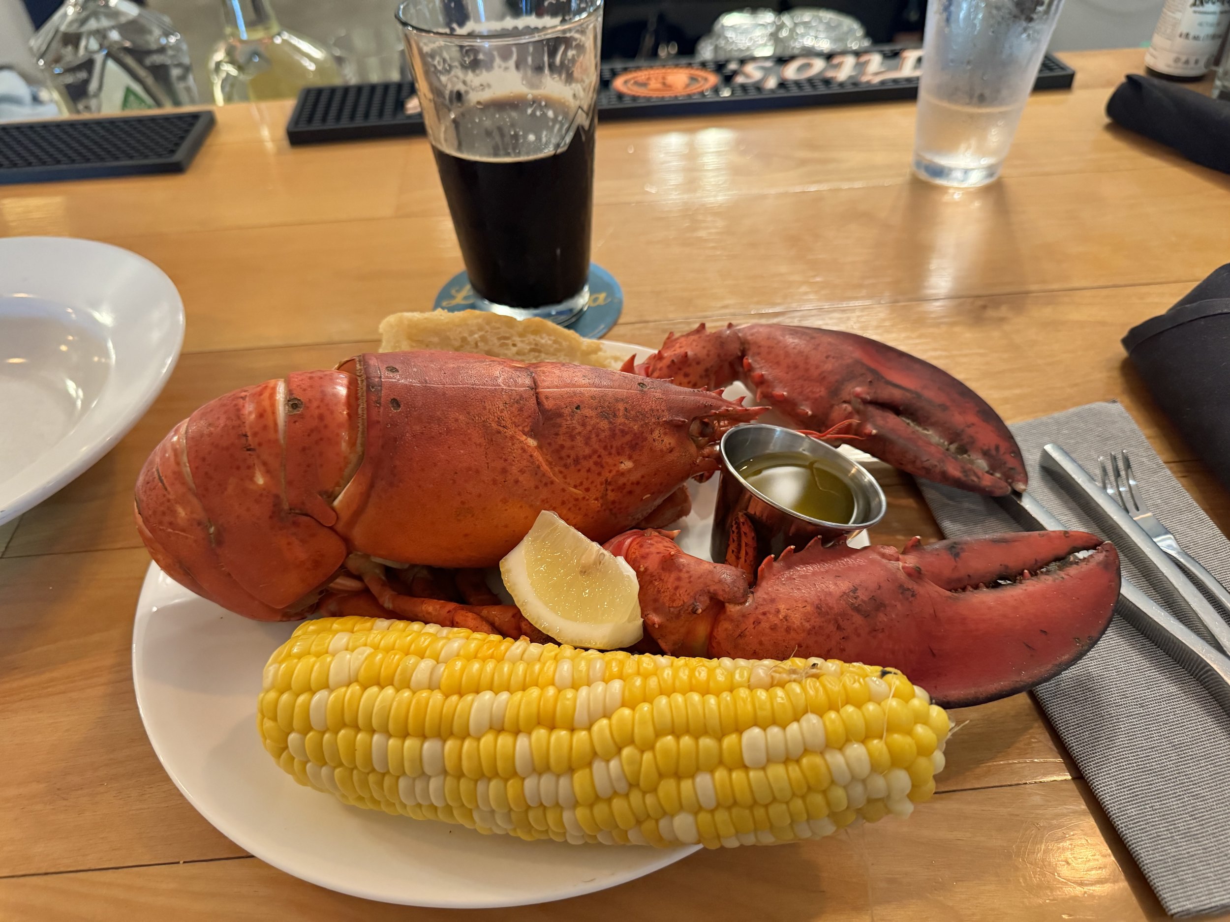 A plate with a cooked lobster, a wedge of lemon, a ear of corn on the cob, and a small metal cup of melted butter. Behind the plate, there is a glass of dark beer and a glass of water on a wooden table.