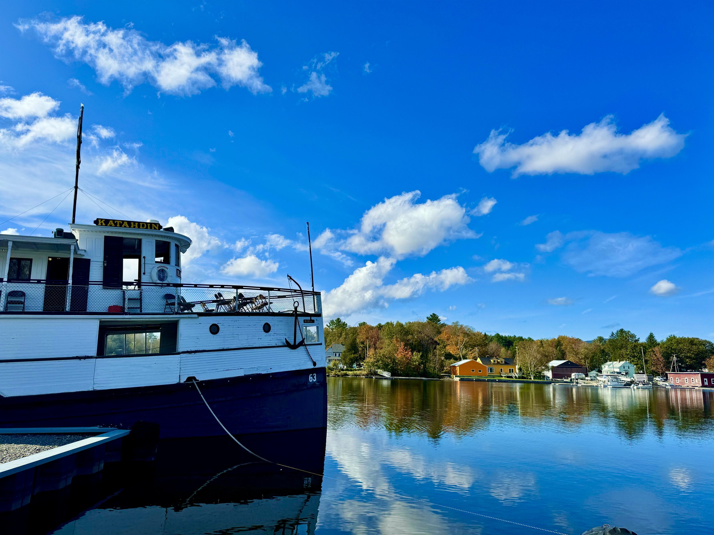 A large white and black boat docked along a calm water body under a partly cloudy blue sky, with charming colorful houses and trees in the background.
