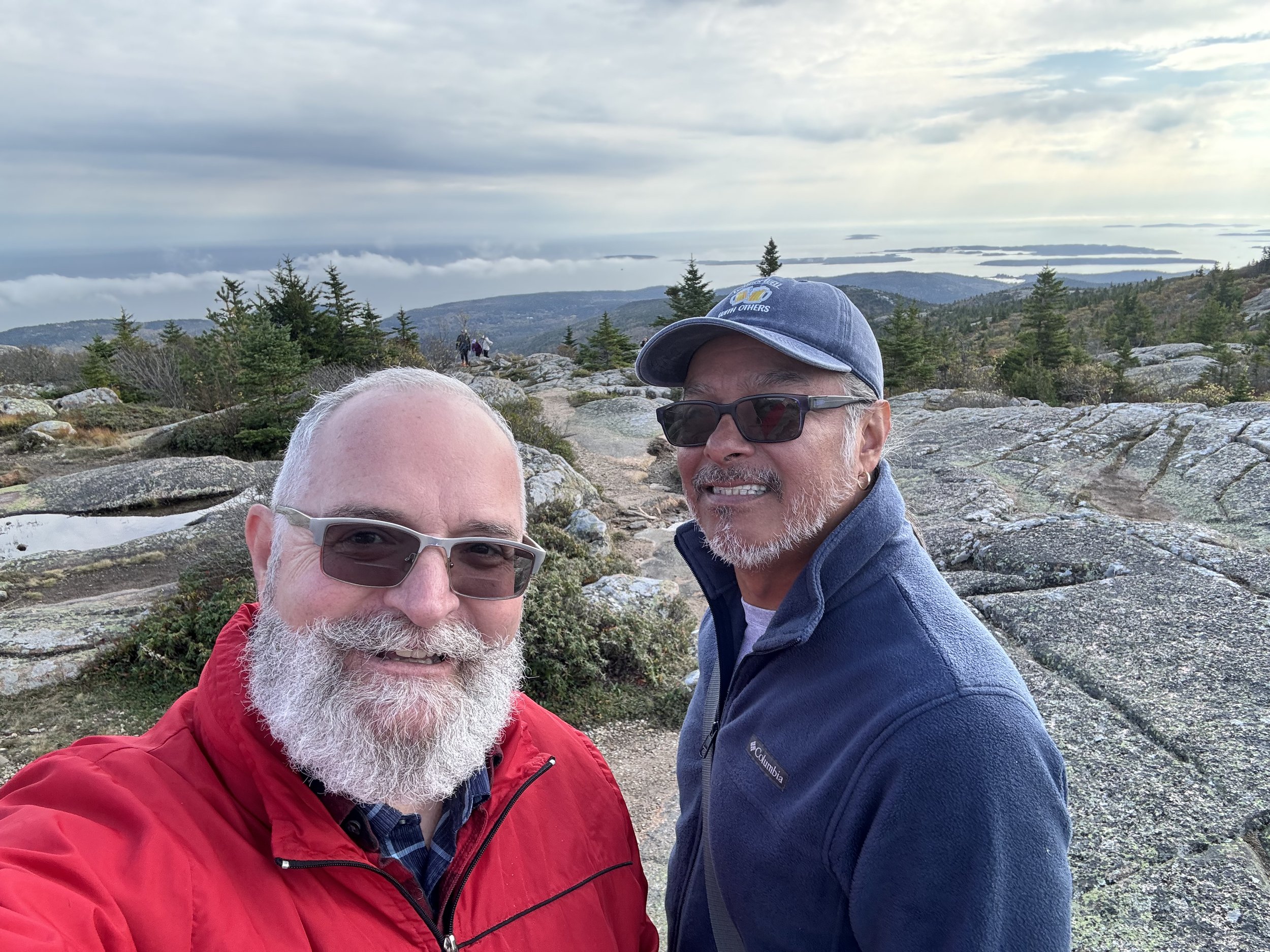 Two men with gray beards wearing sunglasses and outdoor jackets taking a selfie on a mountain trail with a scenic view of trees, rocky terrain, and distant water bodies under an overcast sky.