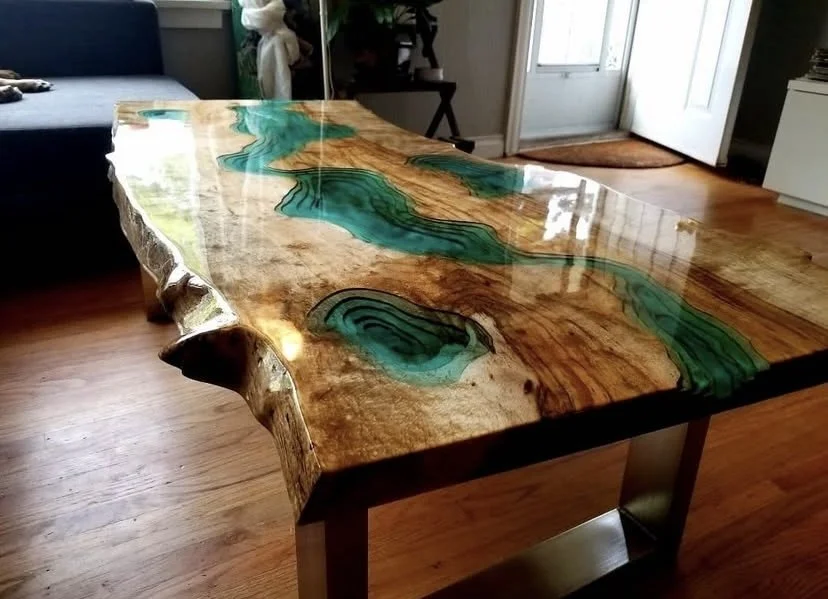 Wooden table with a polished surface and natural edge, featuring green resin inlays highlighting the wood grain, in a room with hardwood floors and a door in the background.