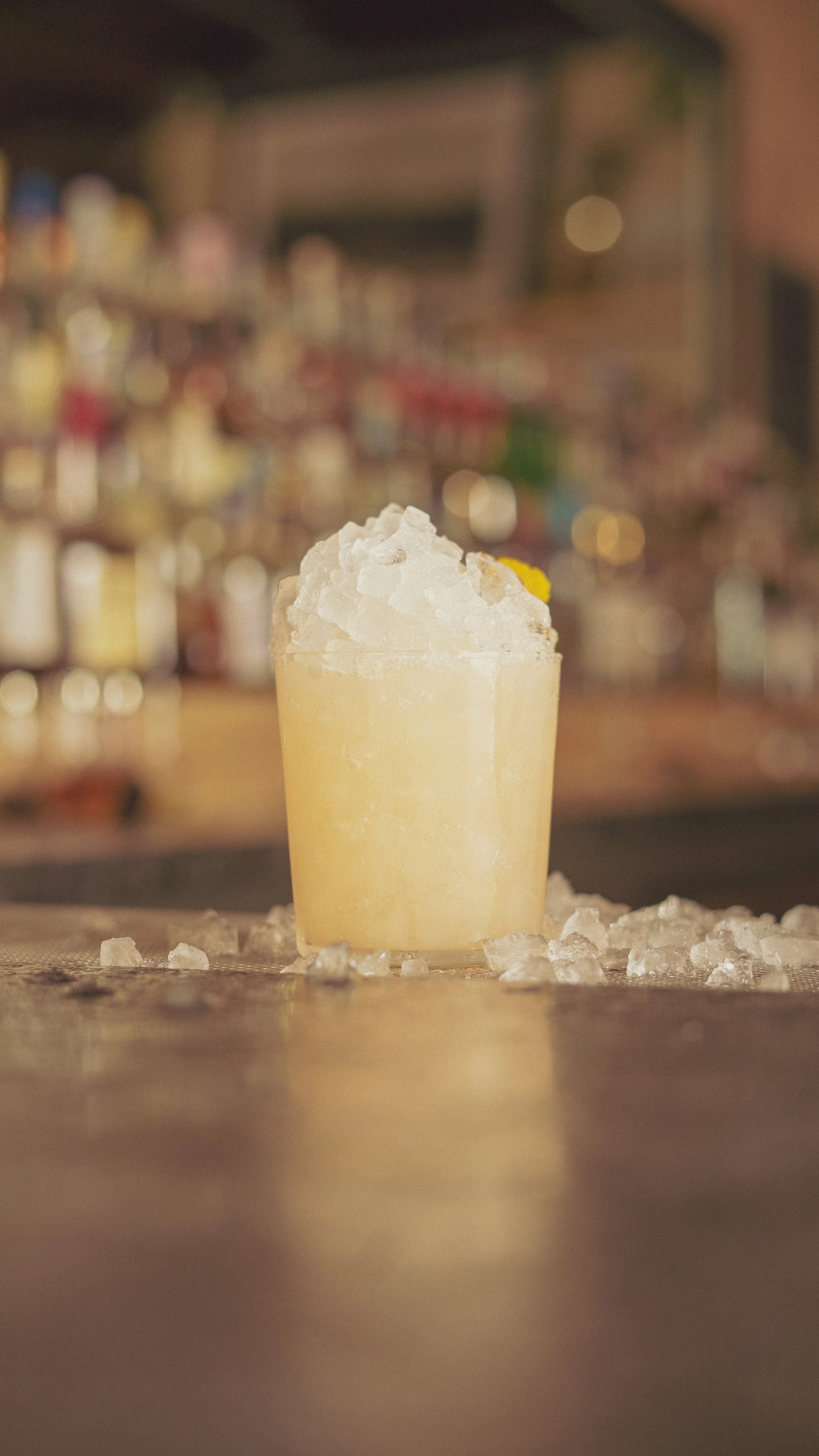 A yellow cocktail garnished with a lemon slice, topped with crushed ice, on a bar counter with scattered ice around it.