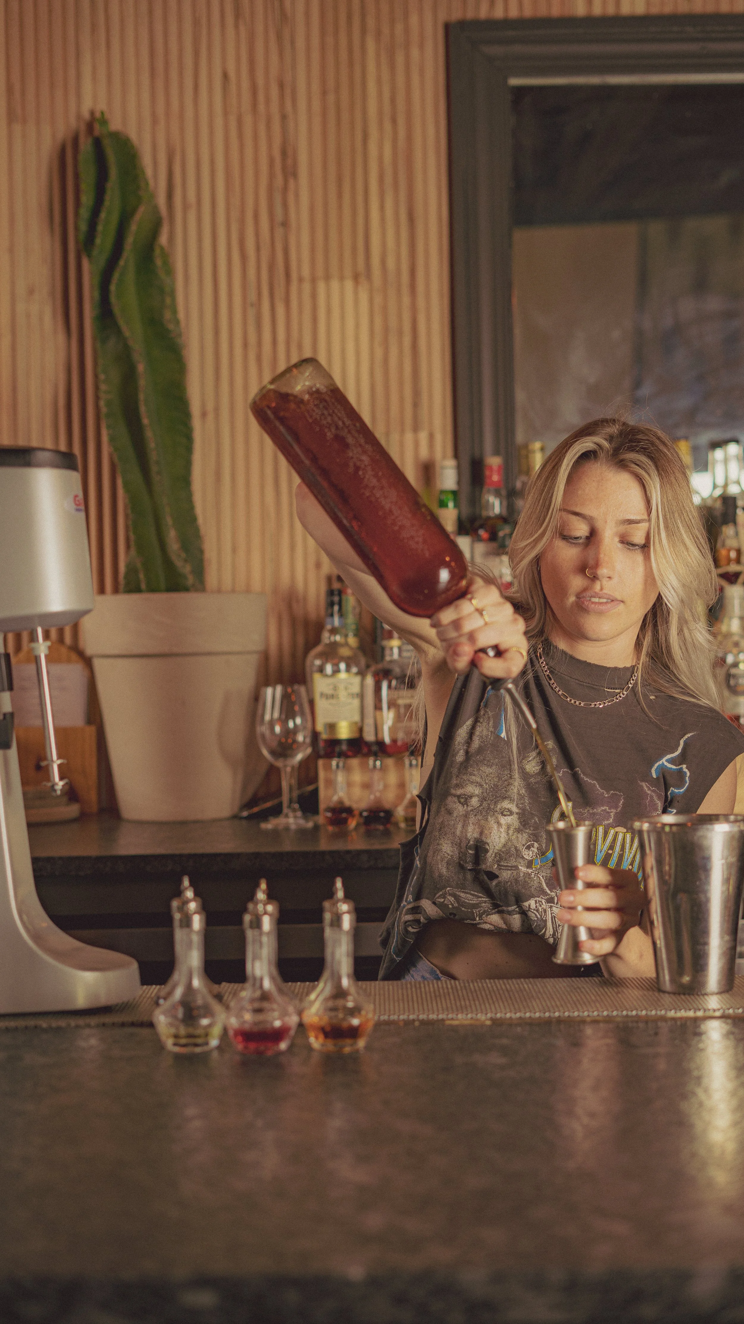 A woman with blonde hair pouring a cocktail into a metal shaker at a bar, with various bottles of alcohol and colorful liquid bottles on the counter in front of her.