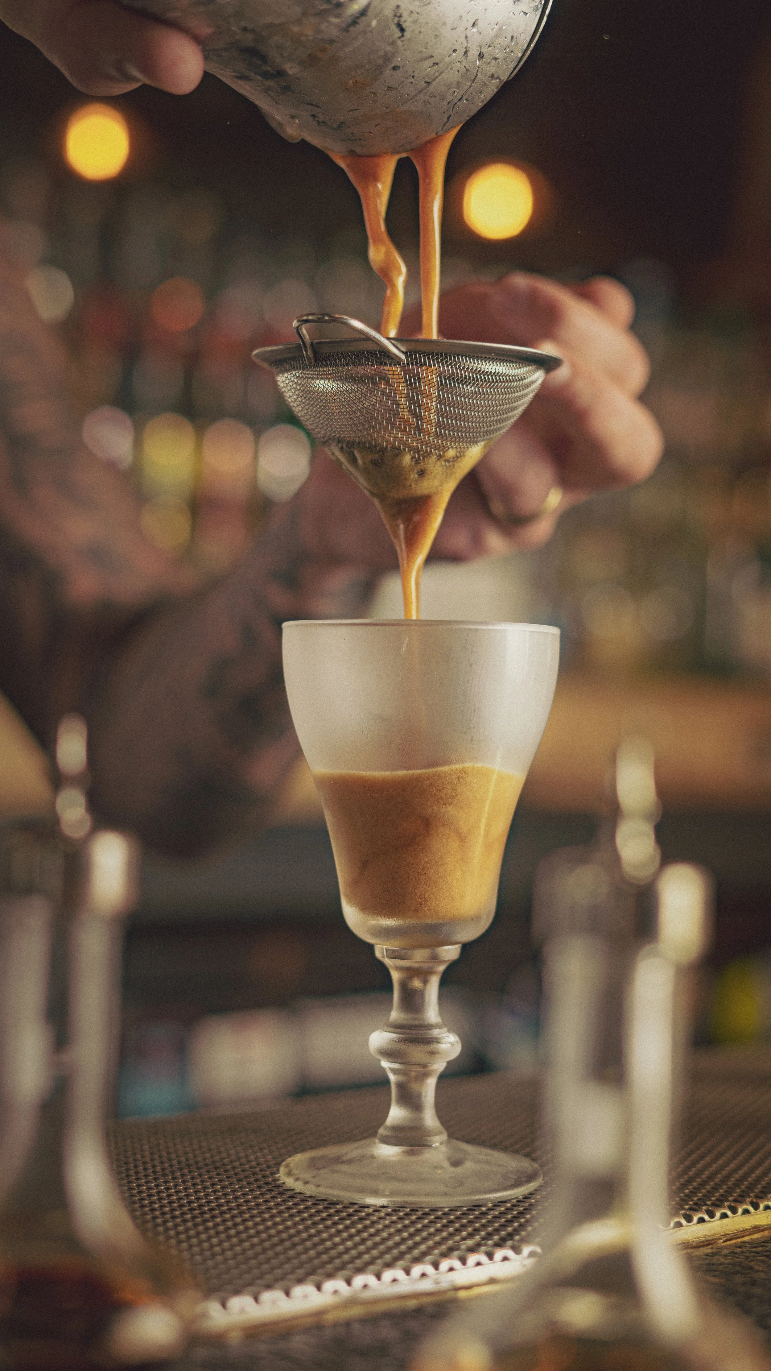 Close-up of a bartender pouring coffee through a strainer into a glass cup layered with ground coffee, creating a layered coffee drink.