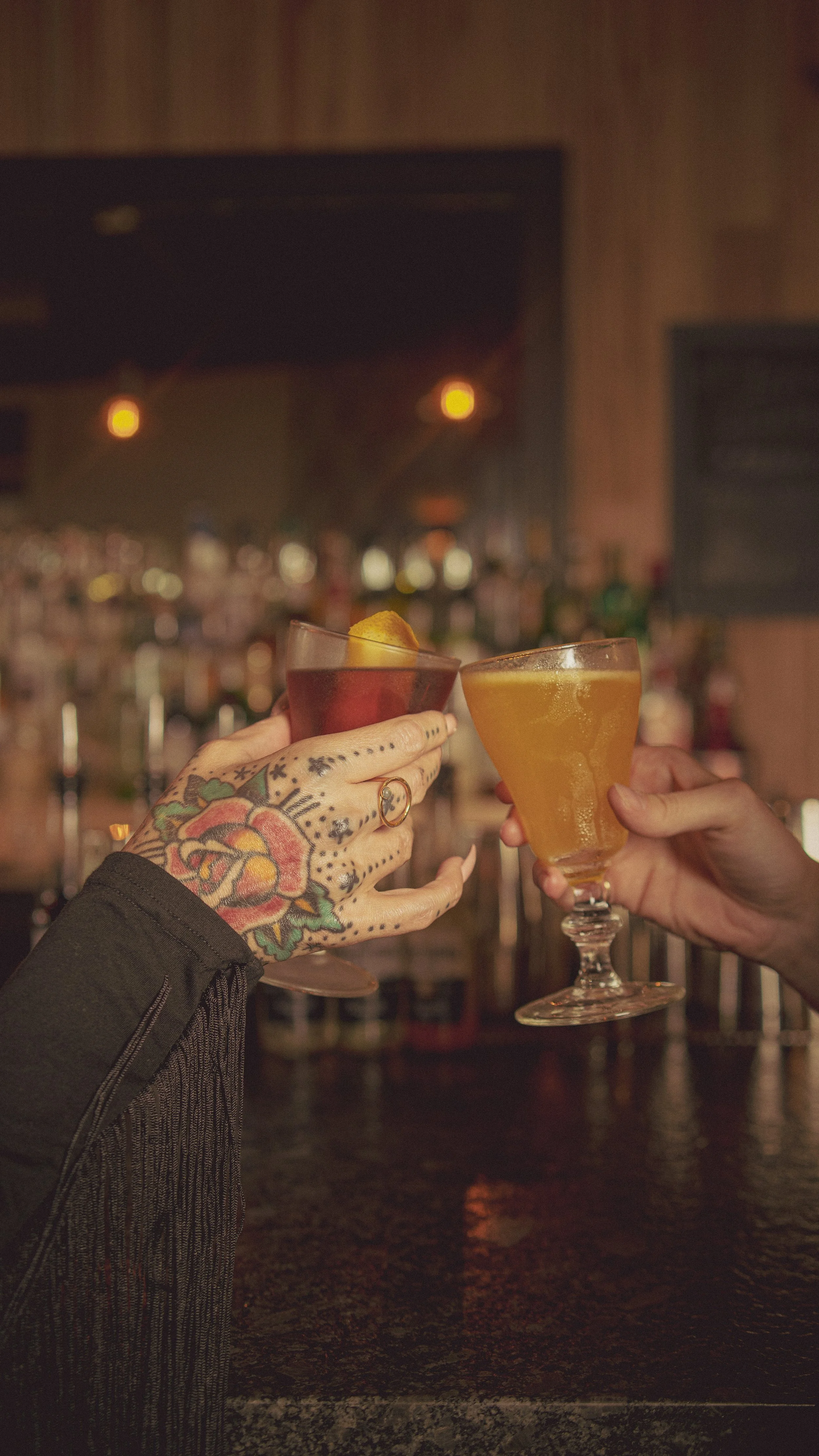 Two people raising cocktails in a toast at a bar, one with a tattooed hand and the other with a plain hand.