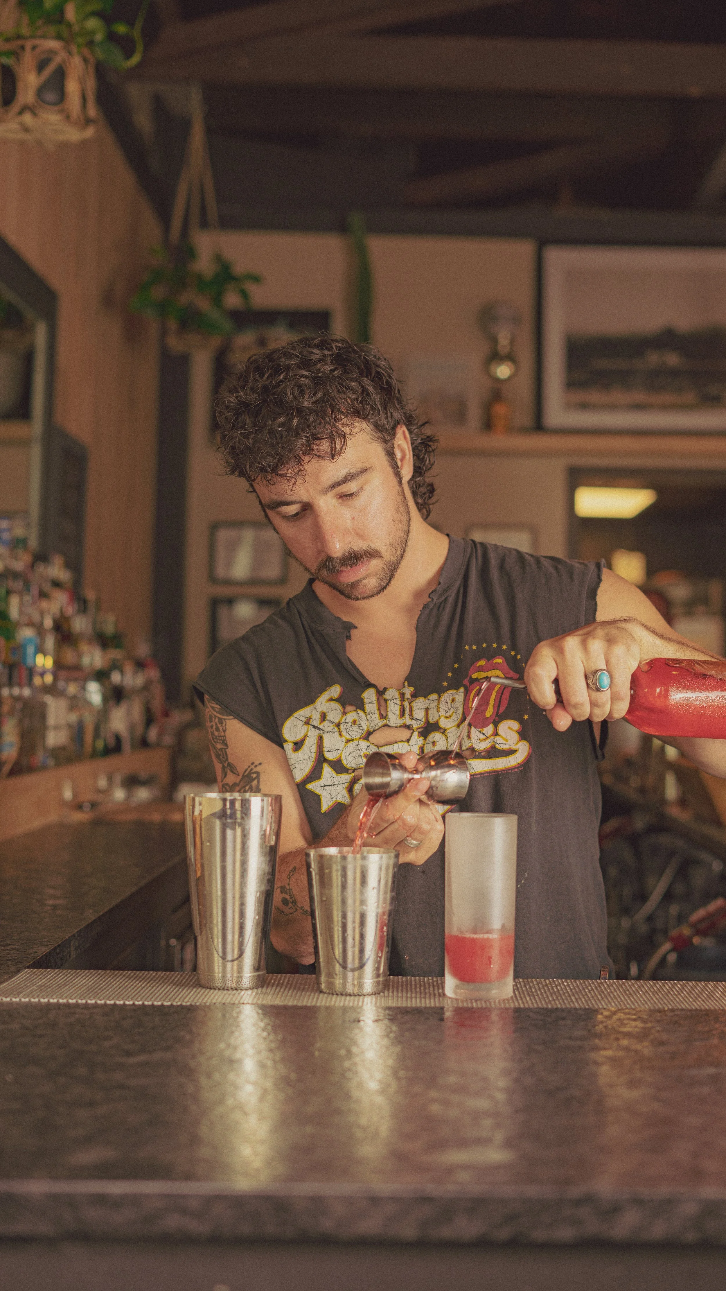 A man with dark curly hair and tattoos on his arm is pouring red liquid into a glass at a bar, wearing a sleeveless Rolling Stones shirt.