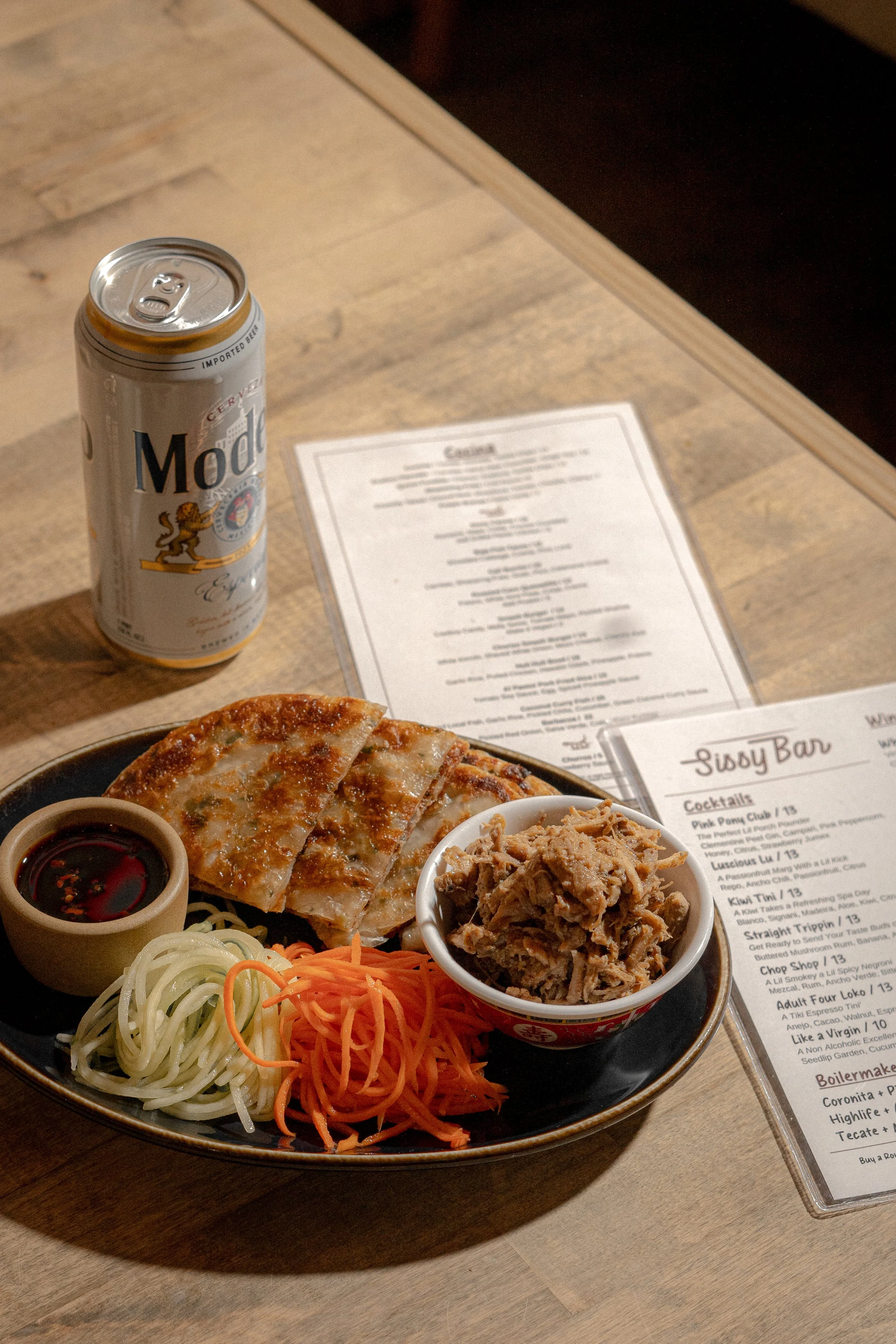 Appetizer plate with gyoza, shredded pork, and vegetables, a can of Modelo beer, and menus on a wooden table.