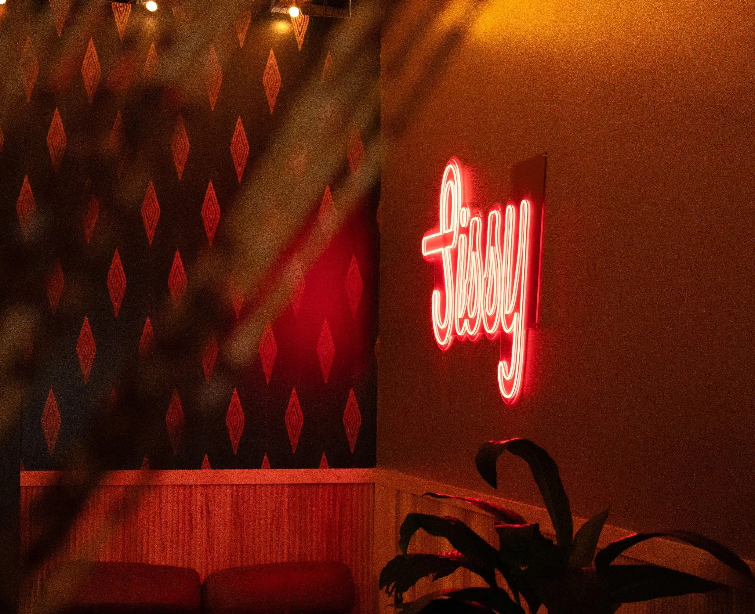 Neon sign reading 'Jewy' in red on a wall with patterned wallpaper, near a plant and seating area.