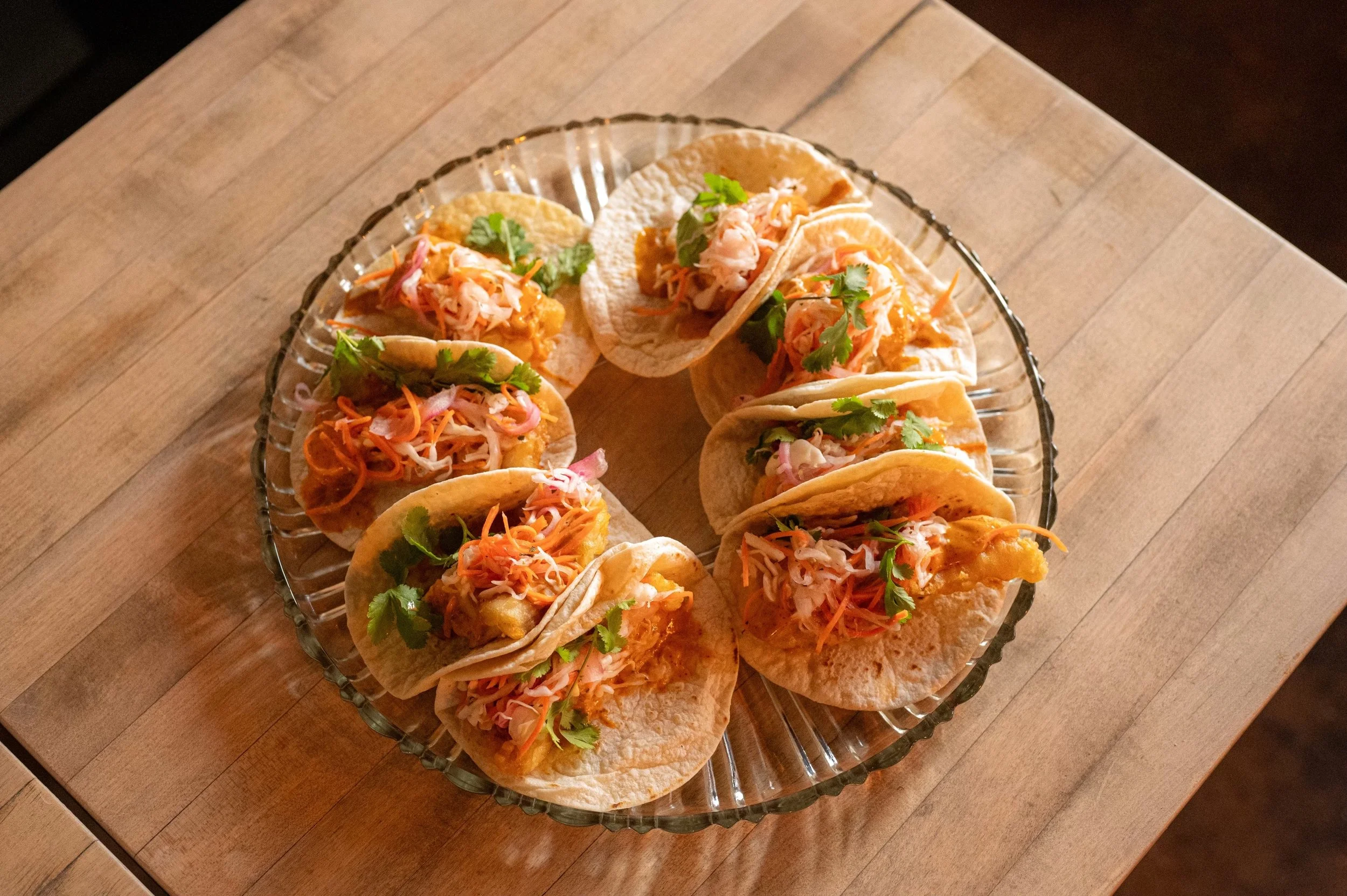 A decorative glass plate holds shrimp tacos topped with cilantro, shredded cabbage, and sliced vegetables, arranged in a circle on a wooden table.