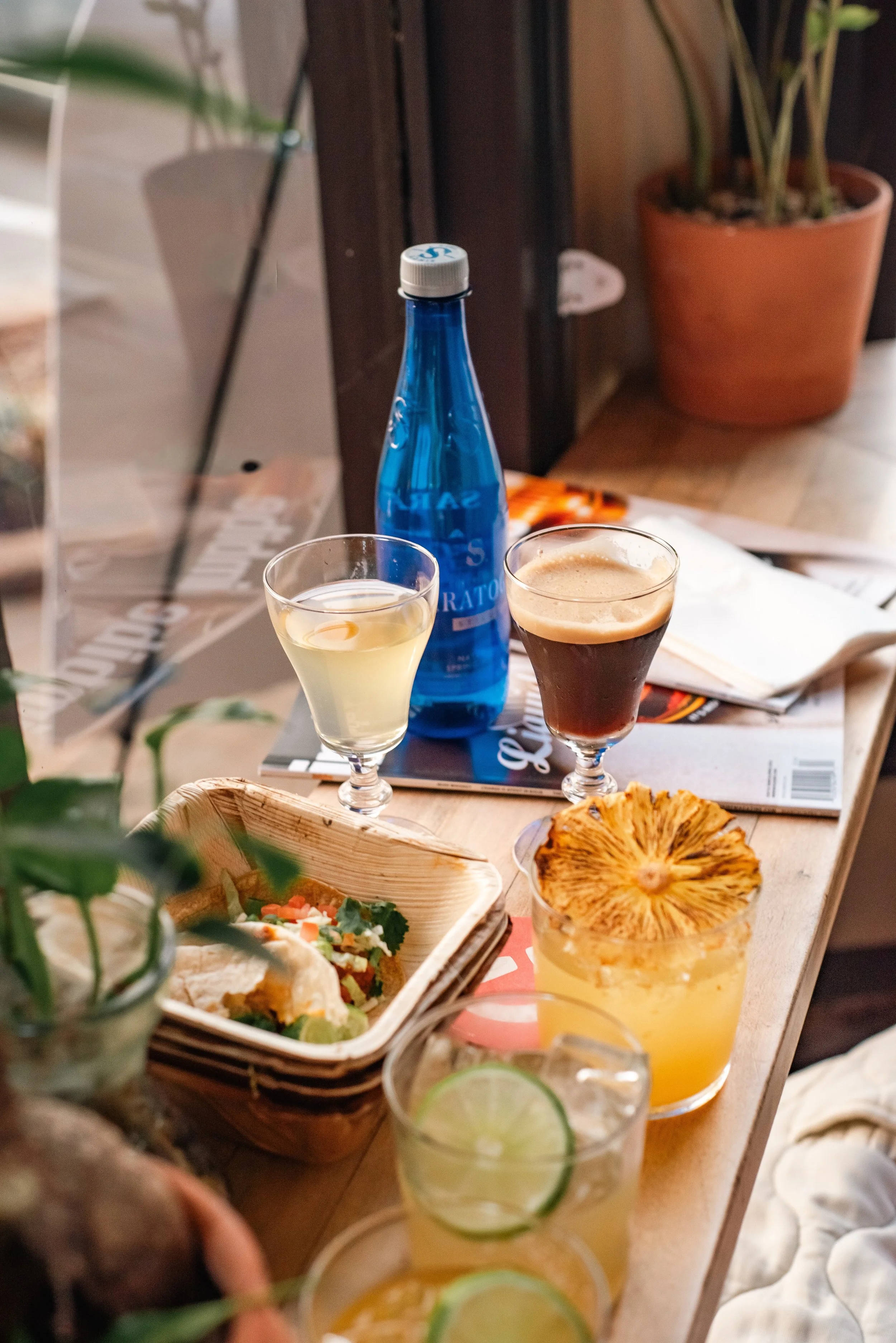 A wooden table with various drinks including a blue water bottle, two glasses of cocktail, a cactus flower, and a taco in a basket. There are also magazines, a notebook, and a plant in the background.