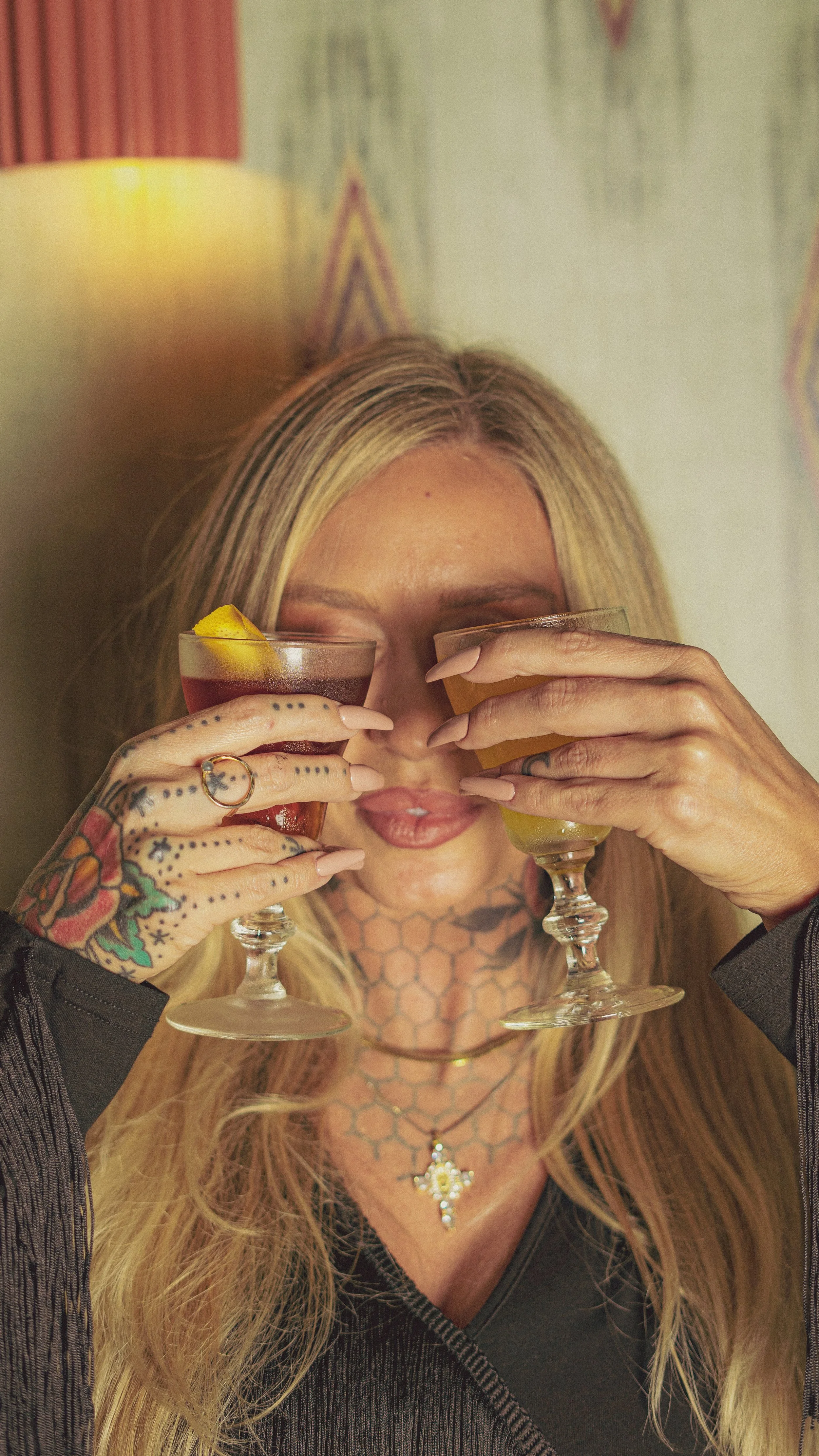 A woman with blonde hair, tattoos, and jewelry holds two glasses with colorful drinks, covering her eyes. She is in a cozy indoor setting.