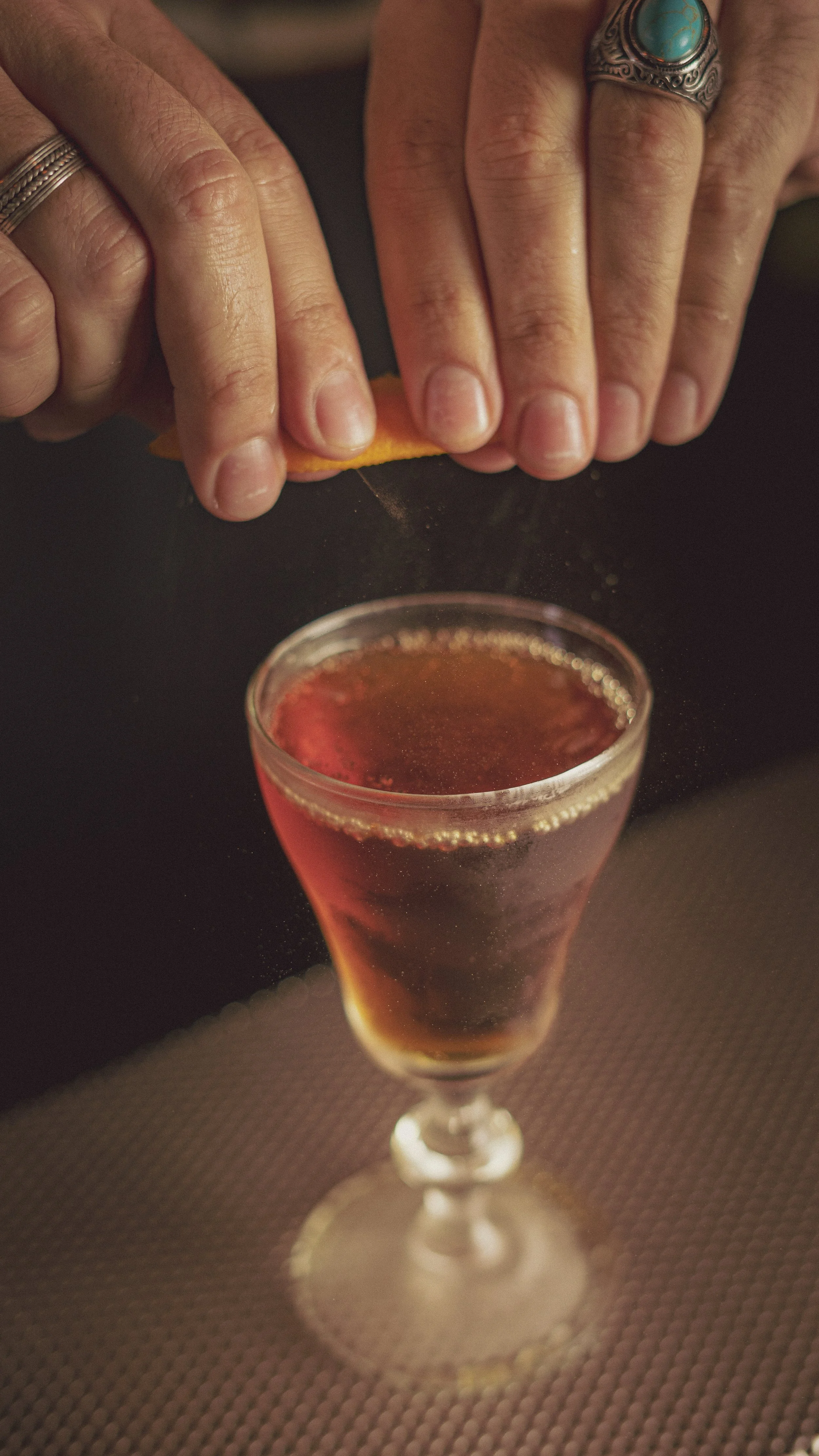 Hands peeling an orange over a glass of dark red liquid, on a dark surface.