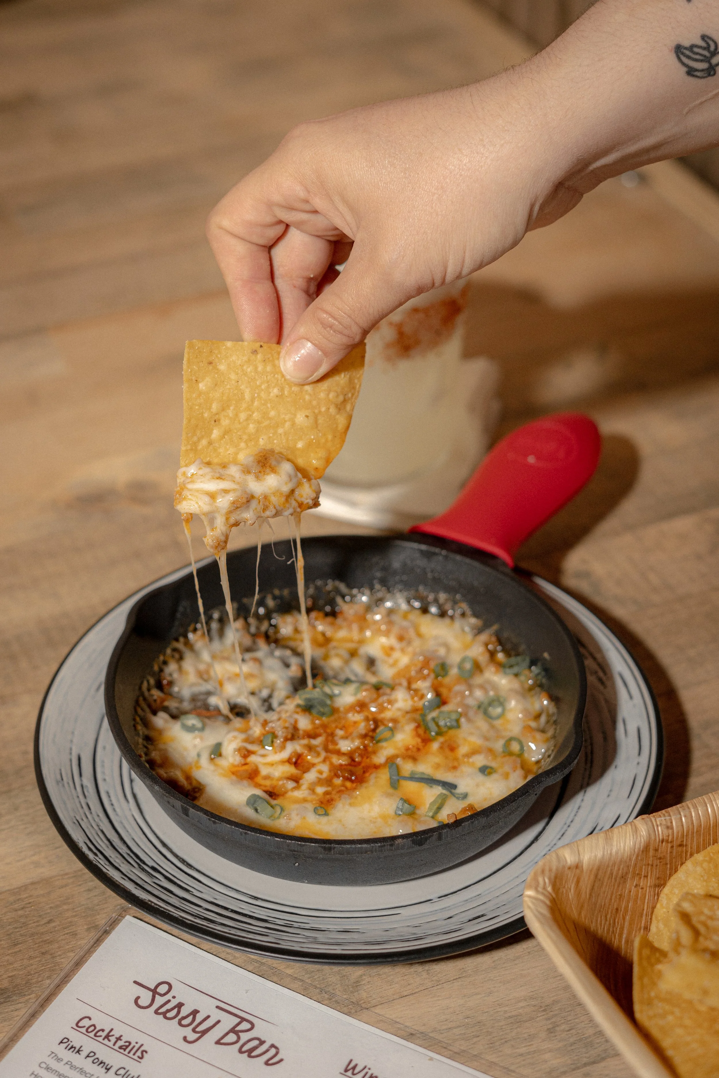 Hand dipping a tortilla chip into a small skillet coated with cheese and topped with chopped green onions.