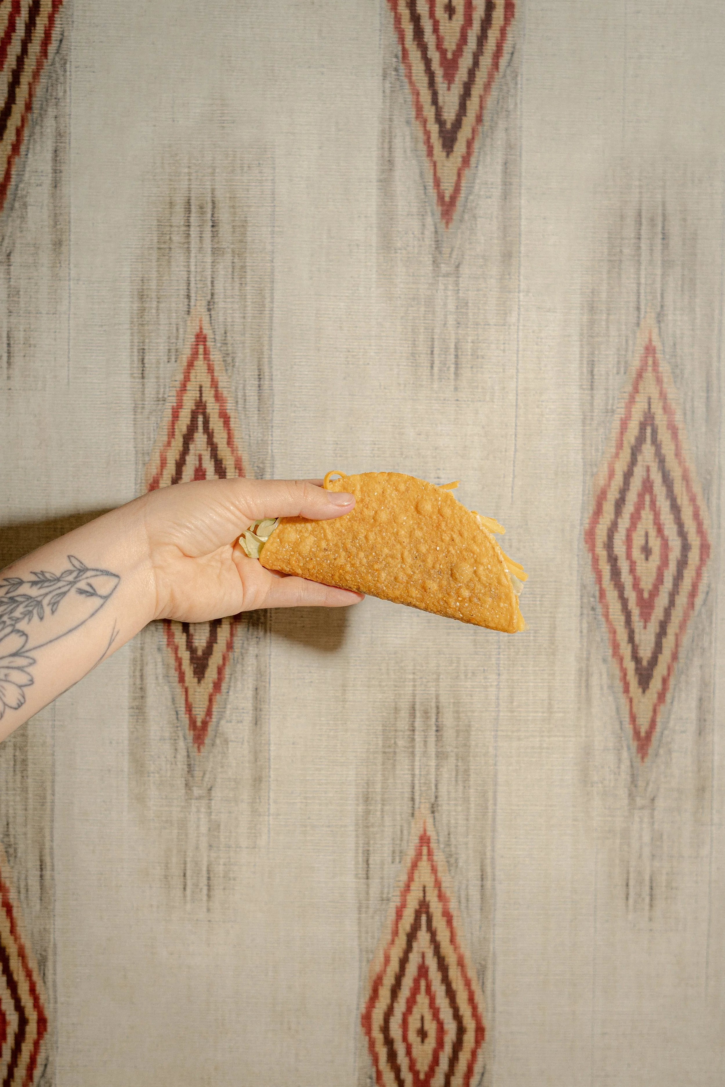 A hand holding a taco with a golden-brown shell, against a patterned fabric background.