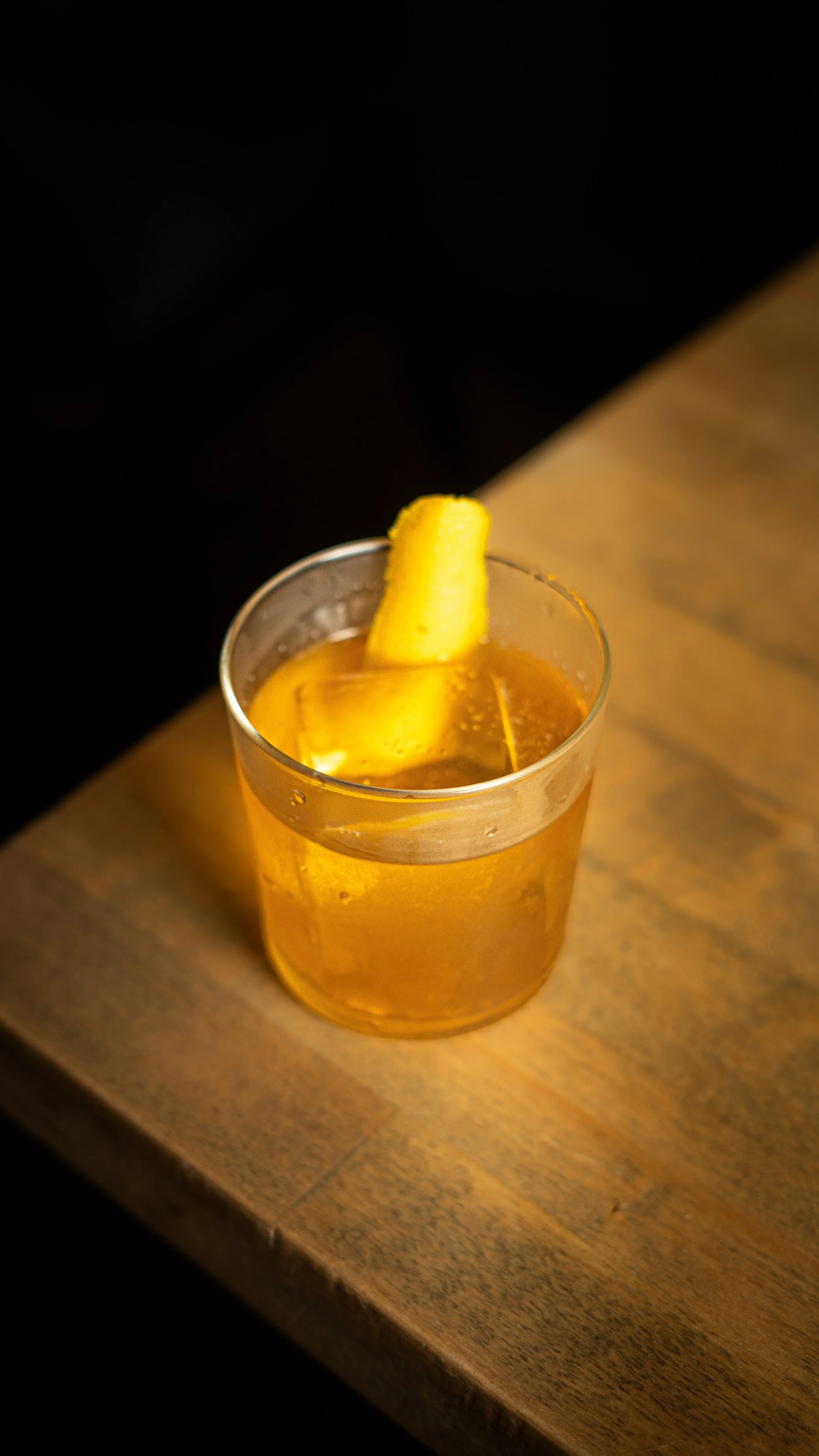 A glass of whiskey with a lemon twist garnish on a wooden table.
