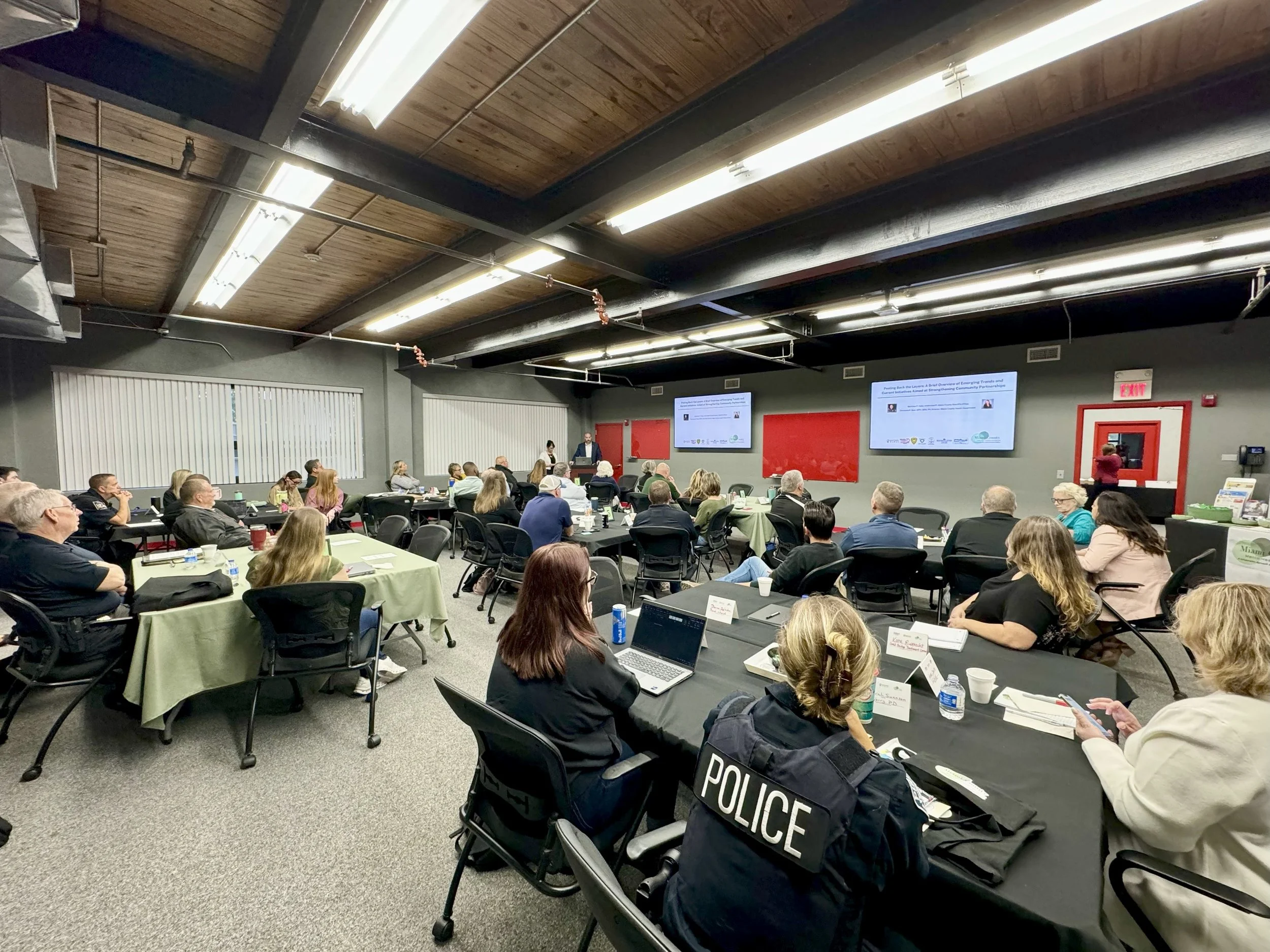 A conference room filled with attendees listening to a presentation on large screens at the front, with some attendees taking notes and a police officer seated at a table.