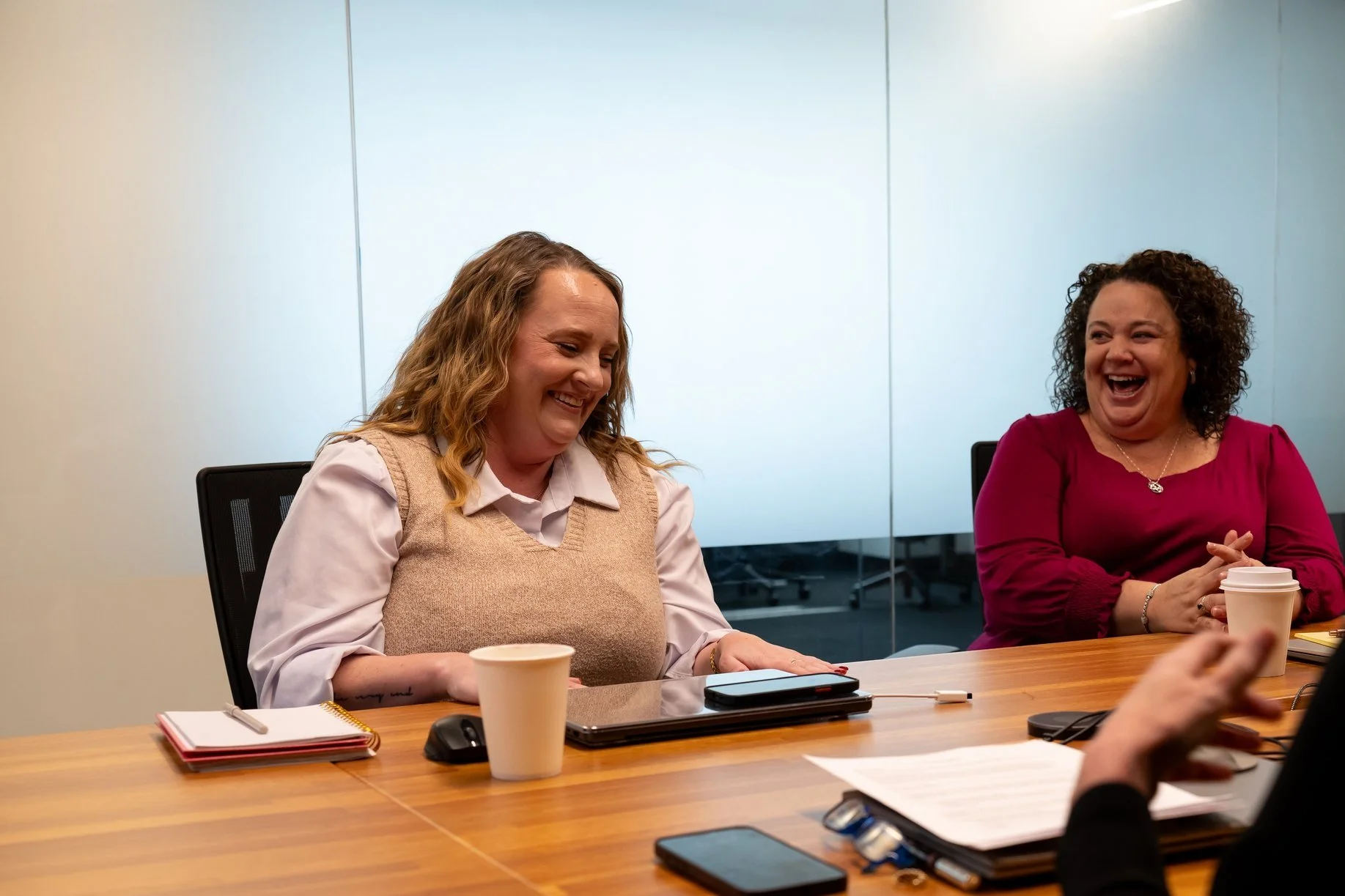 Two women are sitting at a conference table, laughing and engaging in a meeting. One woman wears a beige sweater vest over a white shirt, and the other wears a dark pink top. There are notebooks, cups, and electronic devices on the table.