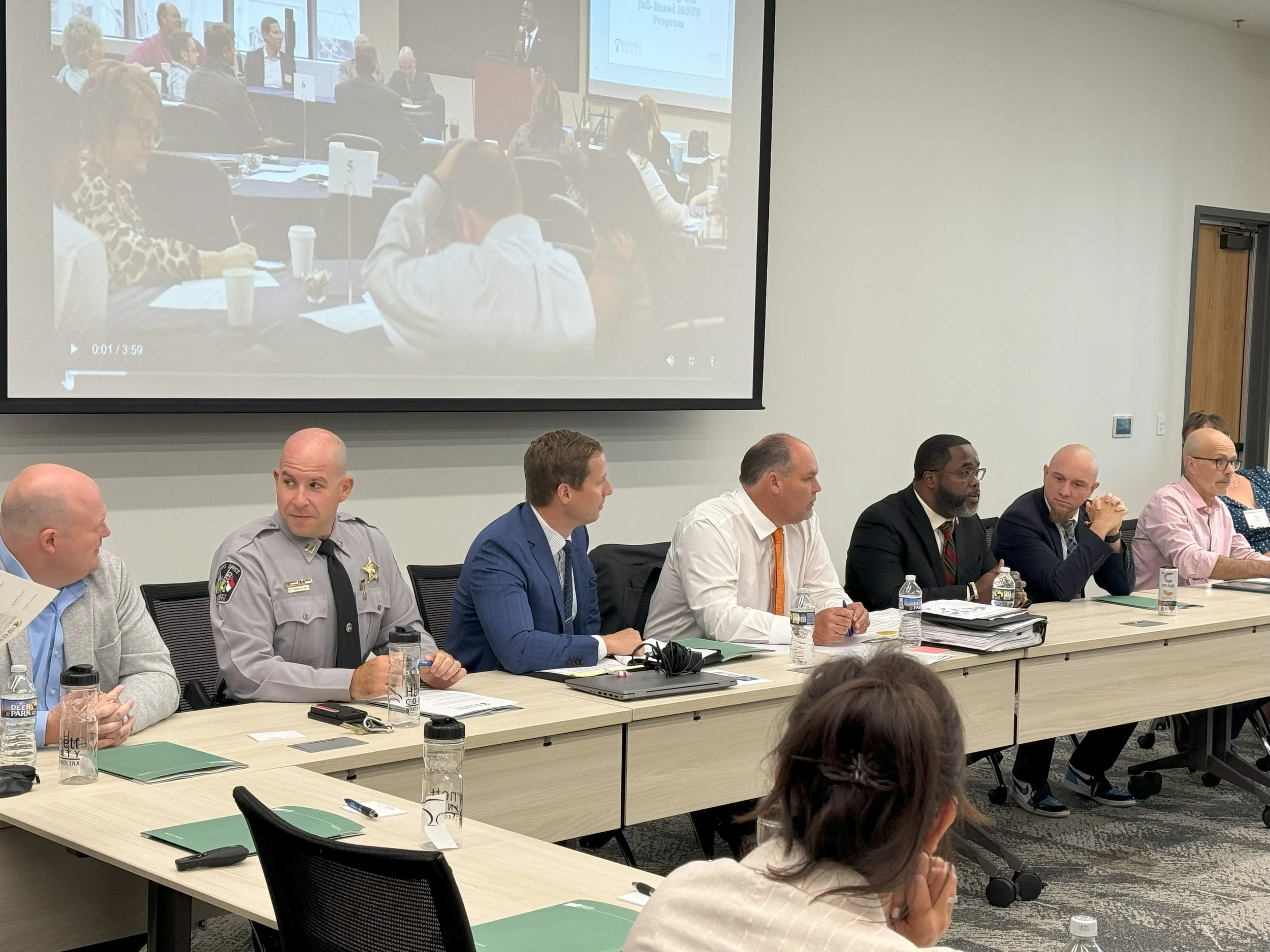 A panel of seven people sitting at a conference table in a meeting room, with a large screen behind them displaying a video of a larger audience in another room.
