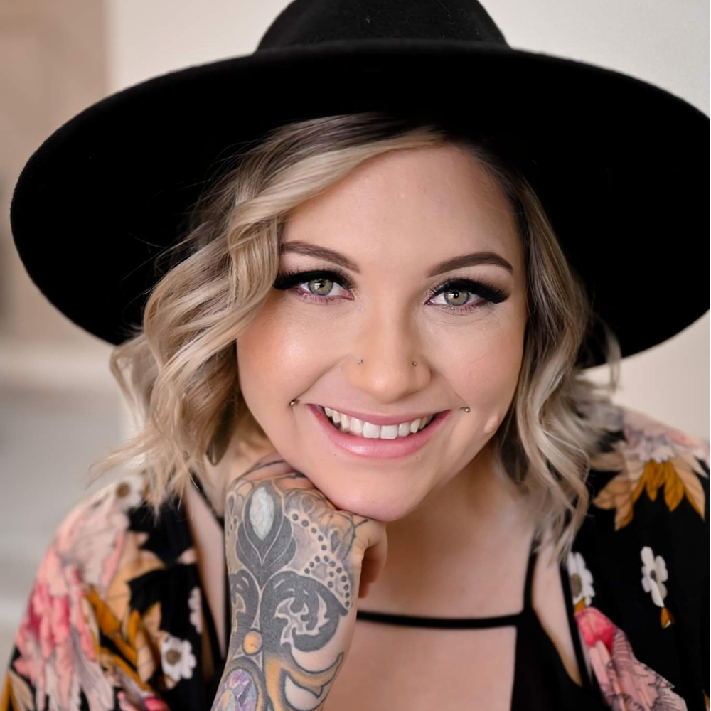 A smiling woman with short blonde hair wearing a black wide-brimmed hat and a floral top.