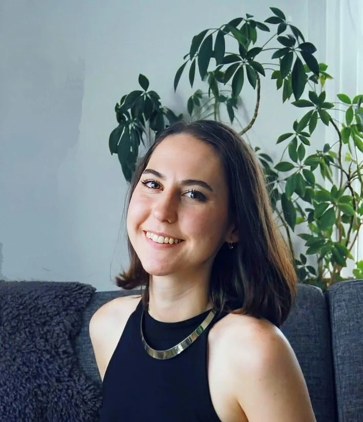 A woman smiling with shoulder-length dark hair, wearing a black sleeveless top and a metallic necklace, sitting on a dark gray couch with a textured throw blanket, with large leafy green houseplants behind her in a well-lit room.