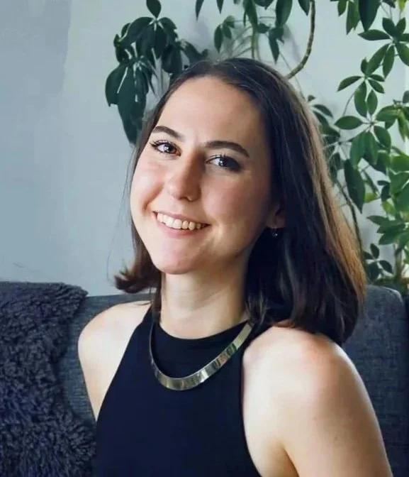 A woman smiling with shoulder-length dark hair, wearing a black sleeveless top and a metallic necklace, sitting on a dark gray couch with a textured throw blanket, with large leafy green houseplants behind her in a well-lit room.