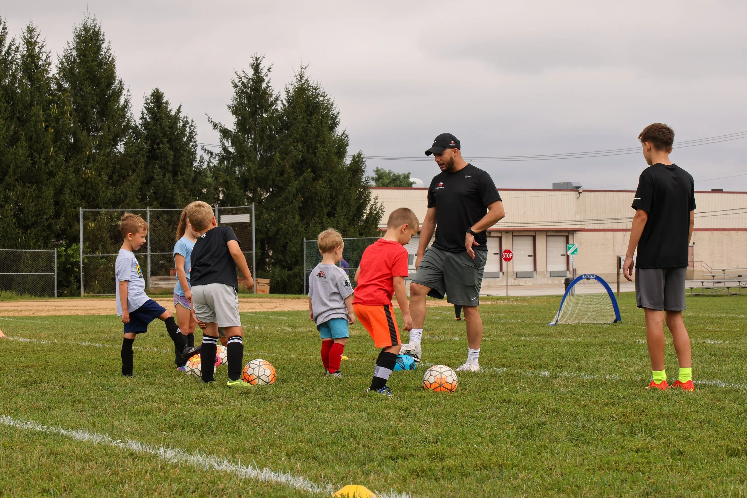 A coach instructs young children during a soccer practice on a grassy field. The children are lined up with soccer balls in front of them, and there are small goal nets in the background.
