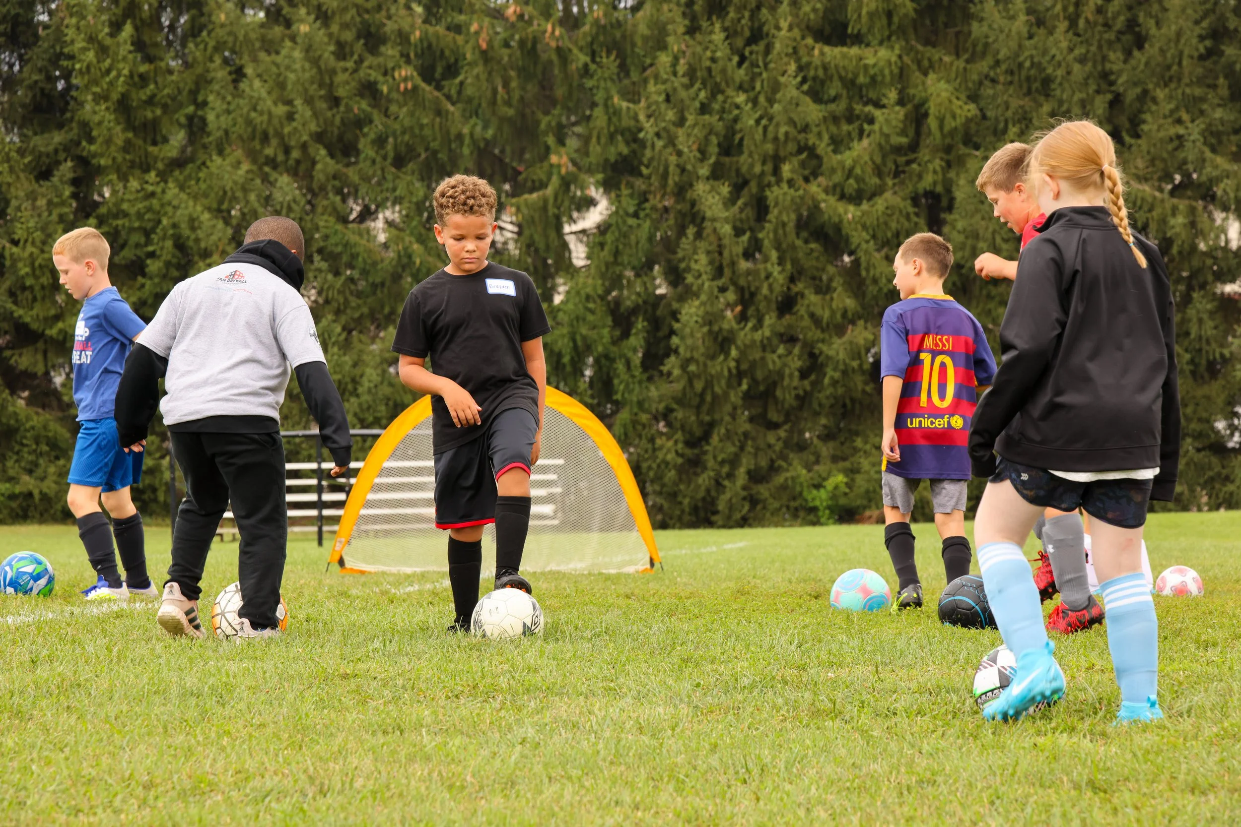 Children practicing soccer on a grassy field with a small goal, wearing colorful sports jerseys and shorts, with trees in the background.