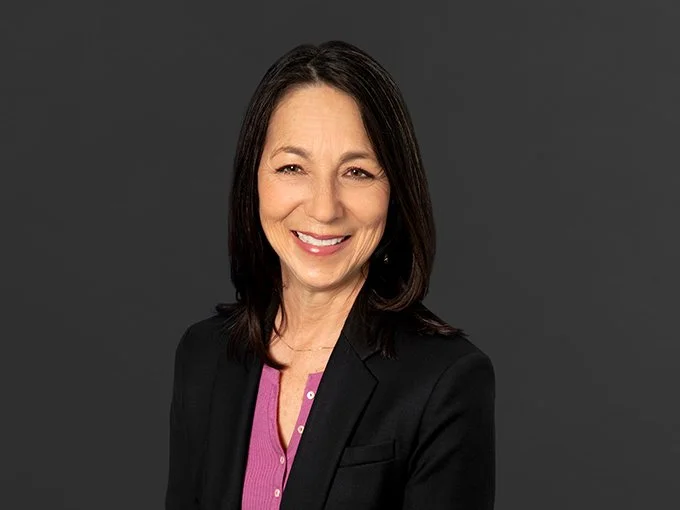 Portrait of a middle-aged woman with shoulder-length dark hair, smiling, wearing a black blazer over a pink blouse, against a gray background.