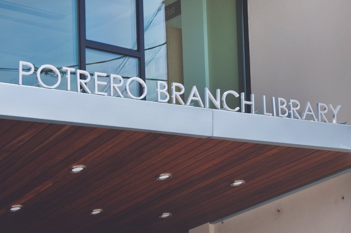 Sign reading 'Potrero Branch Library' on a building exterior with glass windows and a wooden ceiling.