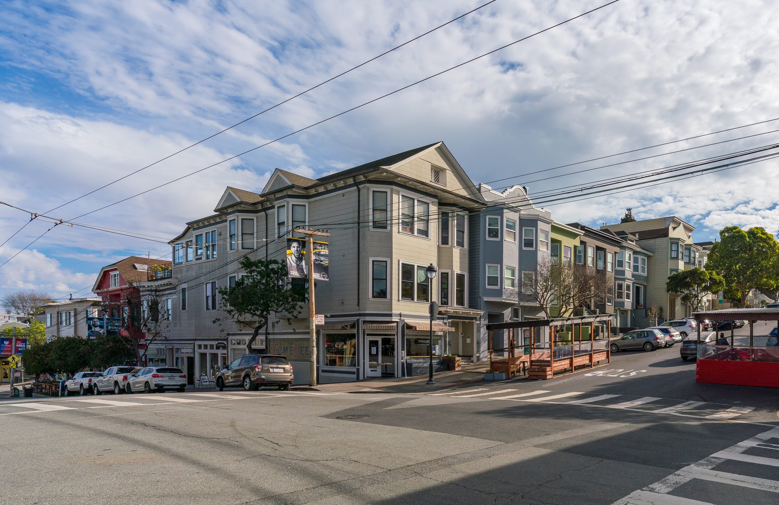 Street view of multi-story residential buildings on a corner, with parked cars, trees, and a partly cloudy sky.