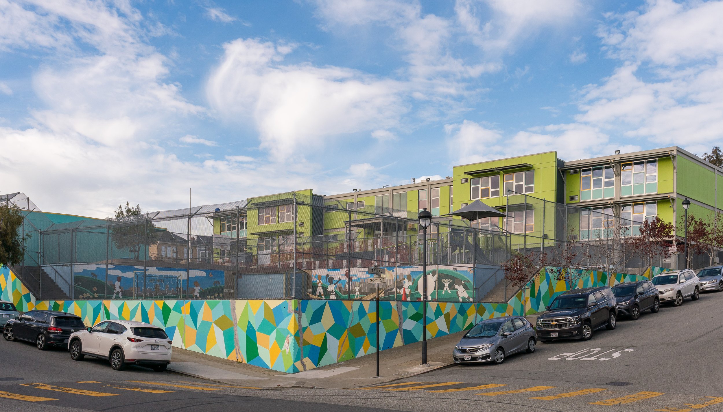 Colorful mural on a fence surrounding a tennis court and modern green apartment buildings on a hill under a partly cloudy sky.